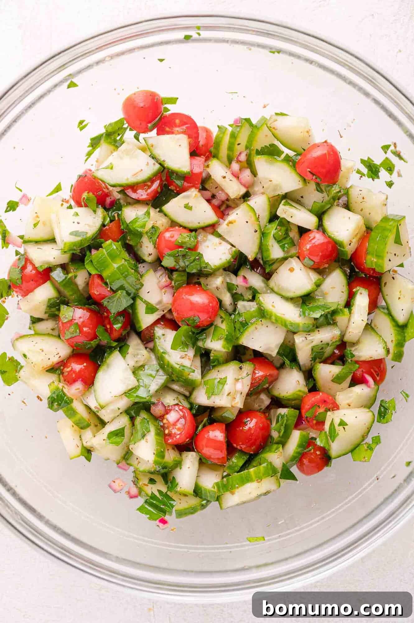 A clear glass bowl showcasing a vibrant mix of chopped cucumbers, juicy cherry tomatoes, finely diced red onions, and fresh parsley, all tossed in a light dressing.