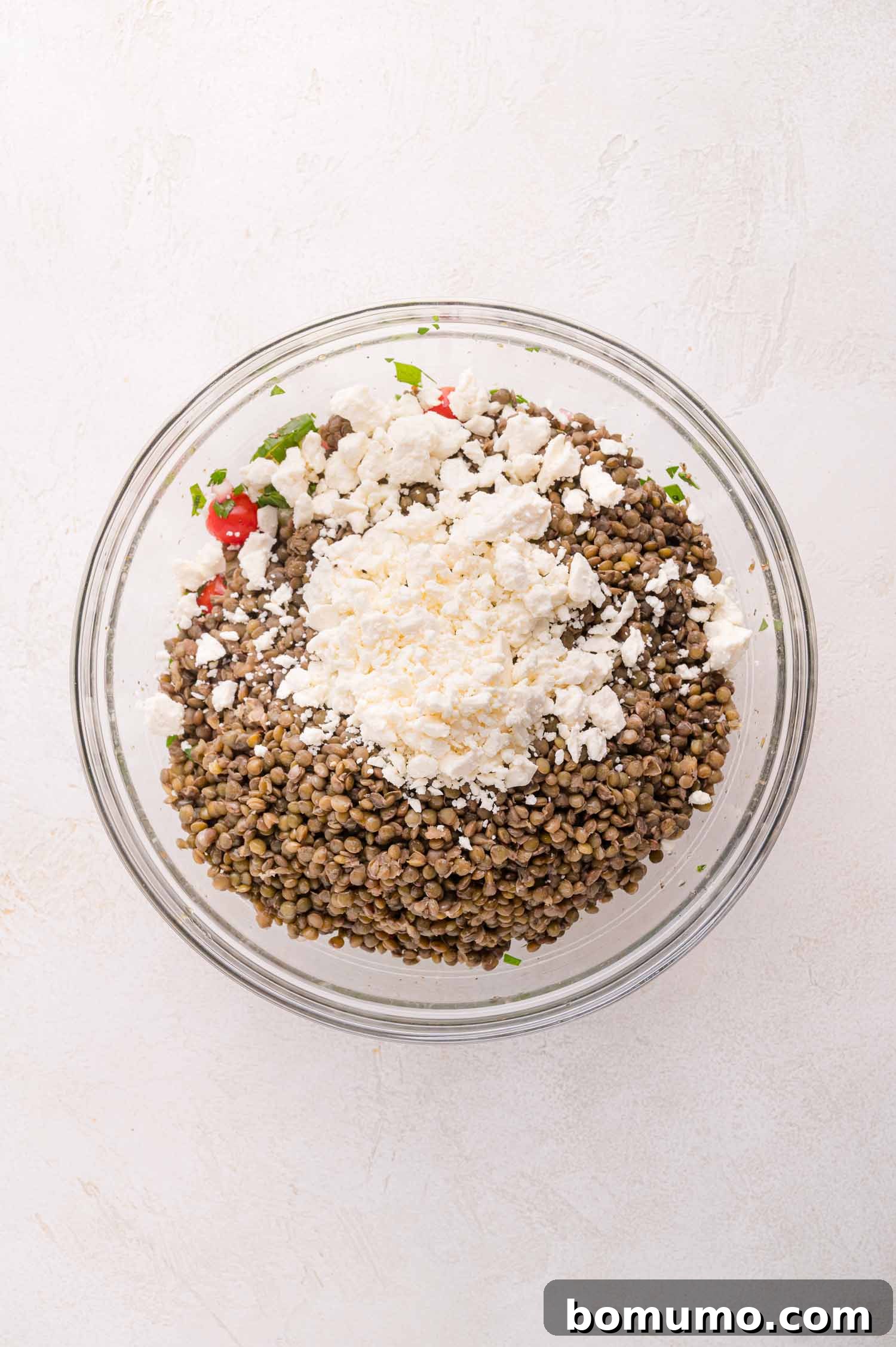 A large glass bowl brimming with a colorful lentil salad, featuring tender lentils, an abundance of chopped vegetables, and a generous topping of creamy crumbled feta cheese, all set on a light-colored surface.