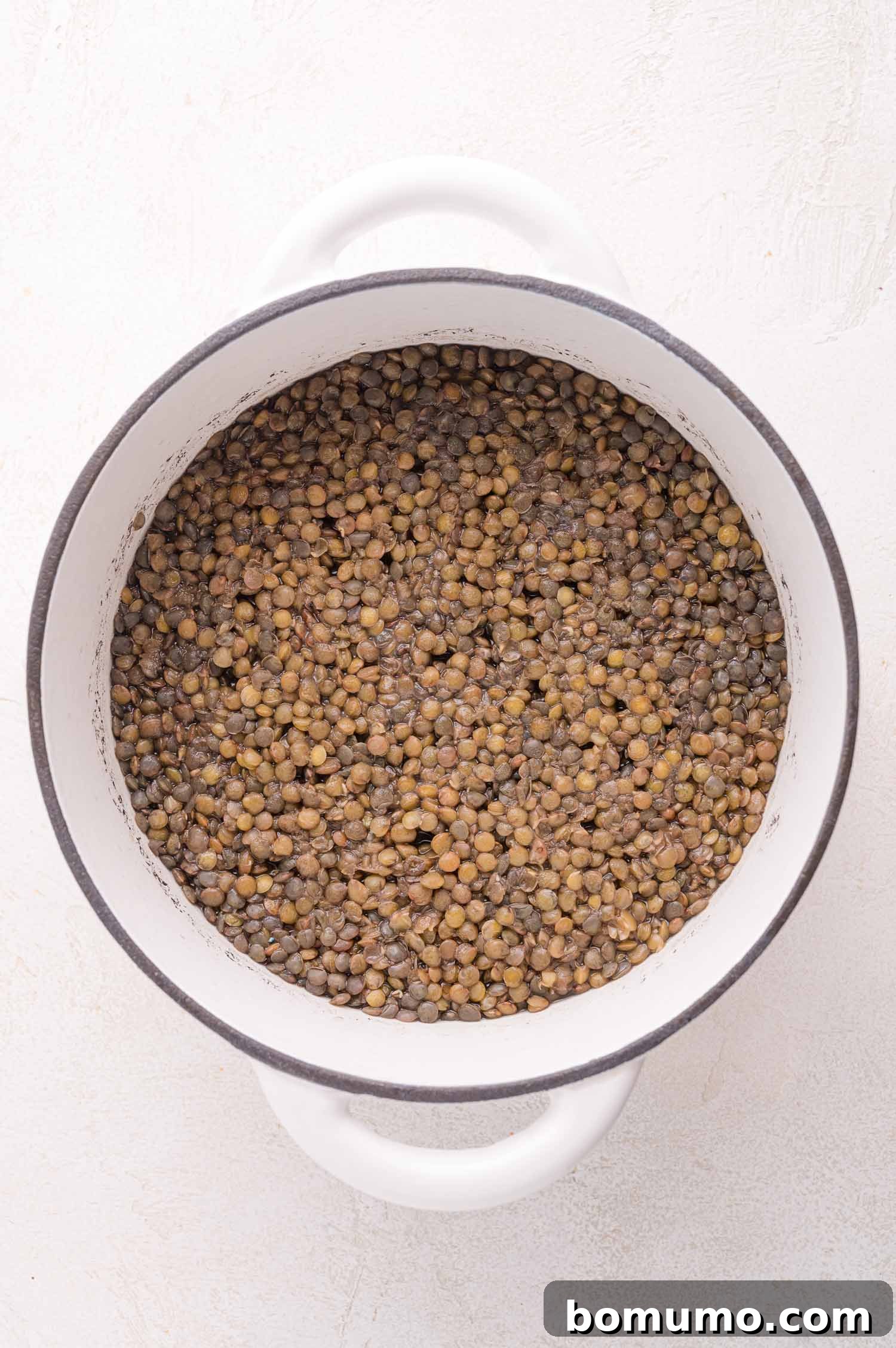 A white pot containing perfectly cooked, tender brown-green lentils, viewed from above on a clean, light-colored surface, ready to be drained and cooled.