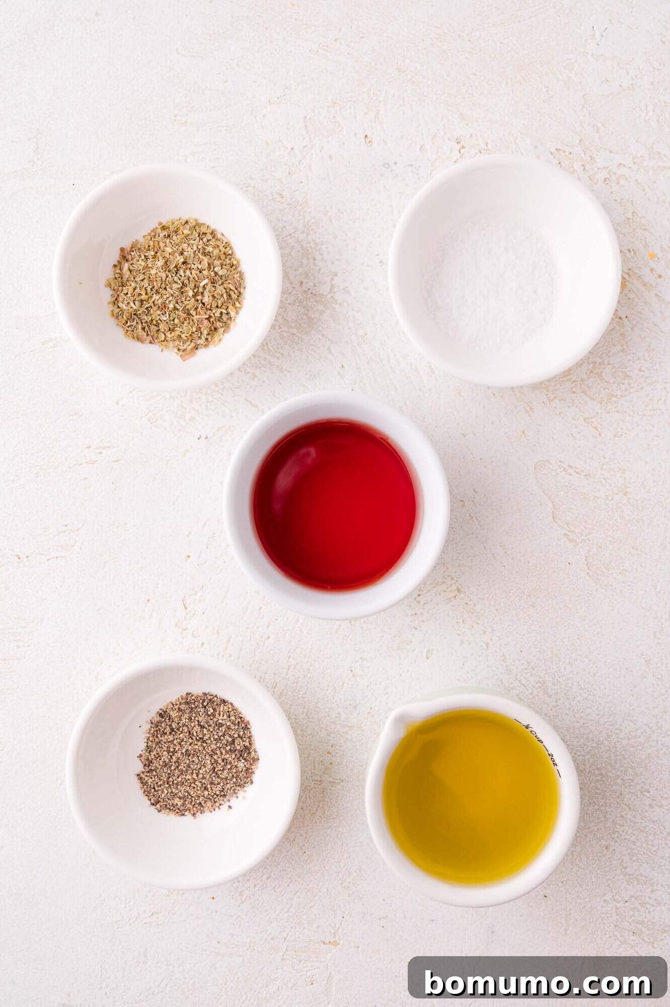 Five small white bowls neatly arranged on a light surface, holding individual ingredients for the vinaigrette: dried oregano, kosher salt, bright red wine vinegar, coarse black pepper, and golden olive oil.