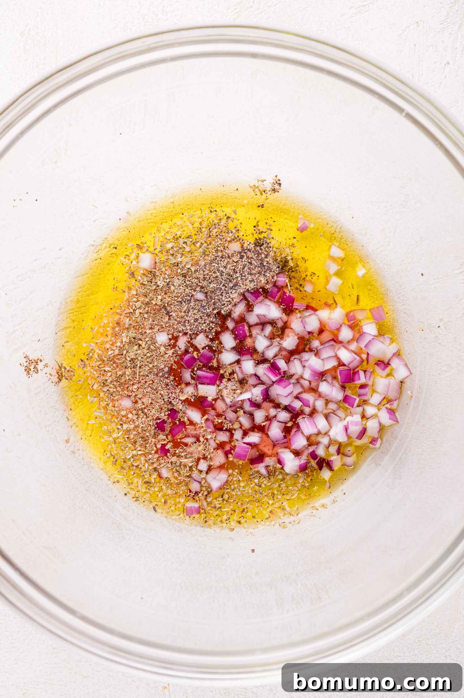A clear glass bowl containing a vibrant mixture of golden olive oil, aromatic dried herbs, and finely chopped red onion, perfectly prepped for the vinaigrette on a light-colored surface.