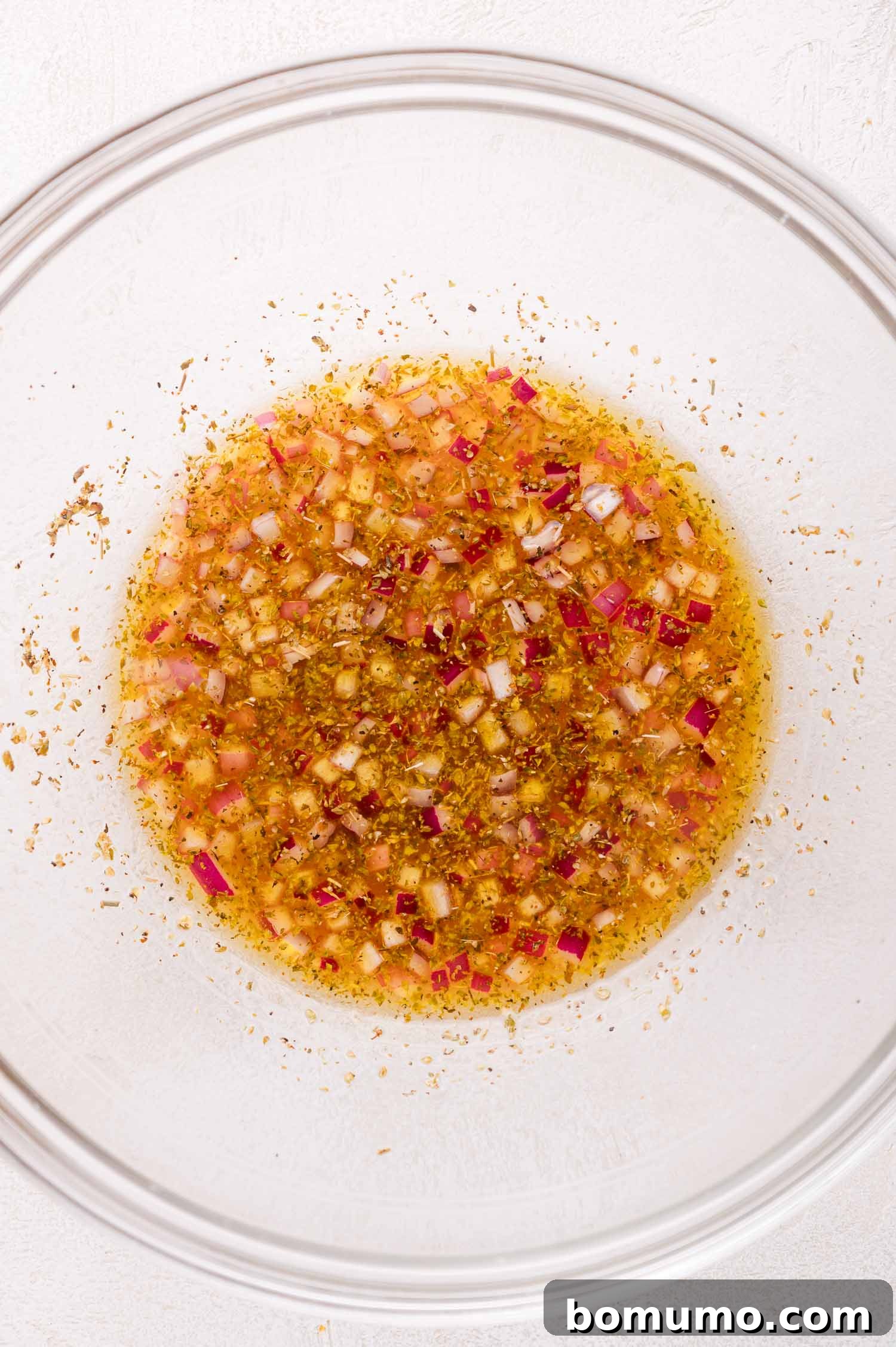 A clear glass bowl showcasing a beautifully emulsified blend of olive oil, red wine vinegar, aromatic red onion, and dried herbs, ready to dress the salad on a crisp white surface.
