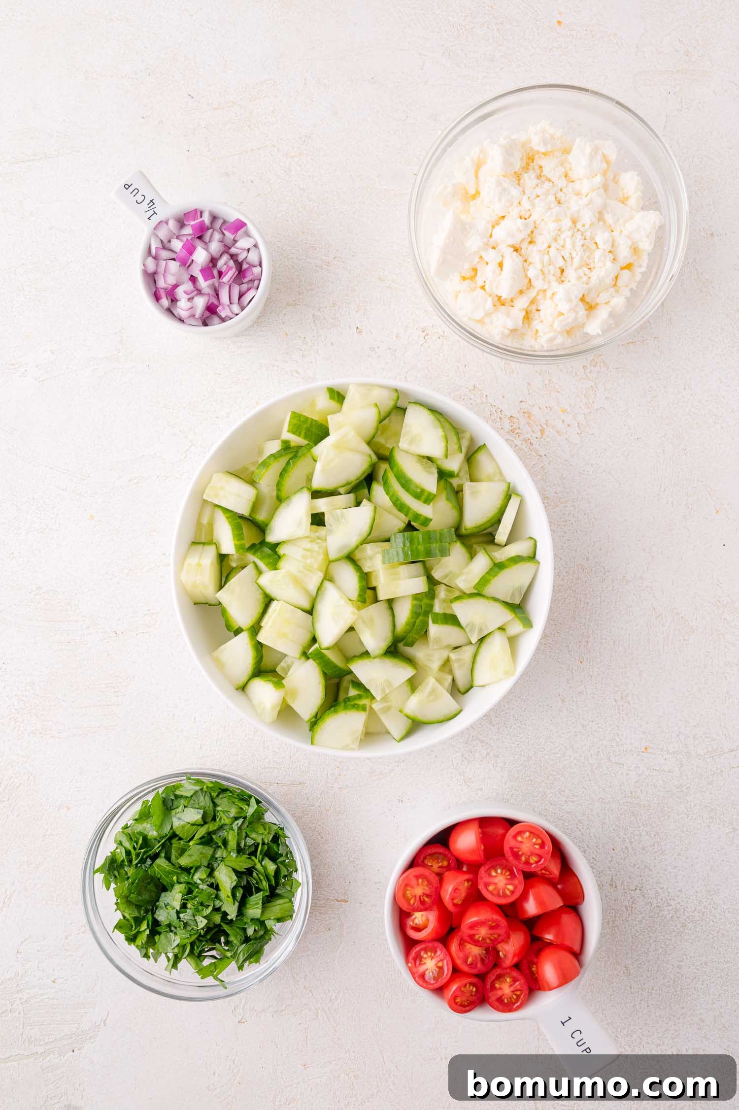 Five brightly colored bowls featuring meticulously prepared ingredients for the lentil salad: chopped red onion, crumbled feta cheese, crisp sliced cucumber, juicy halved cherry tomatoes, and aromatic chopped parsley, all arranged on a light surface.