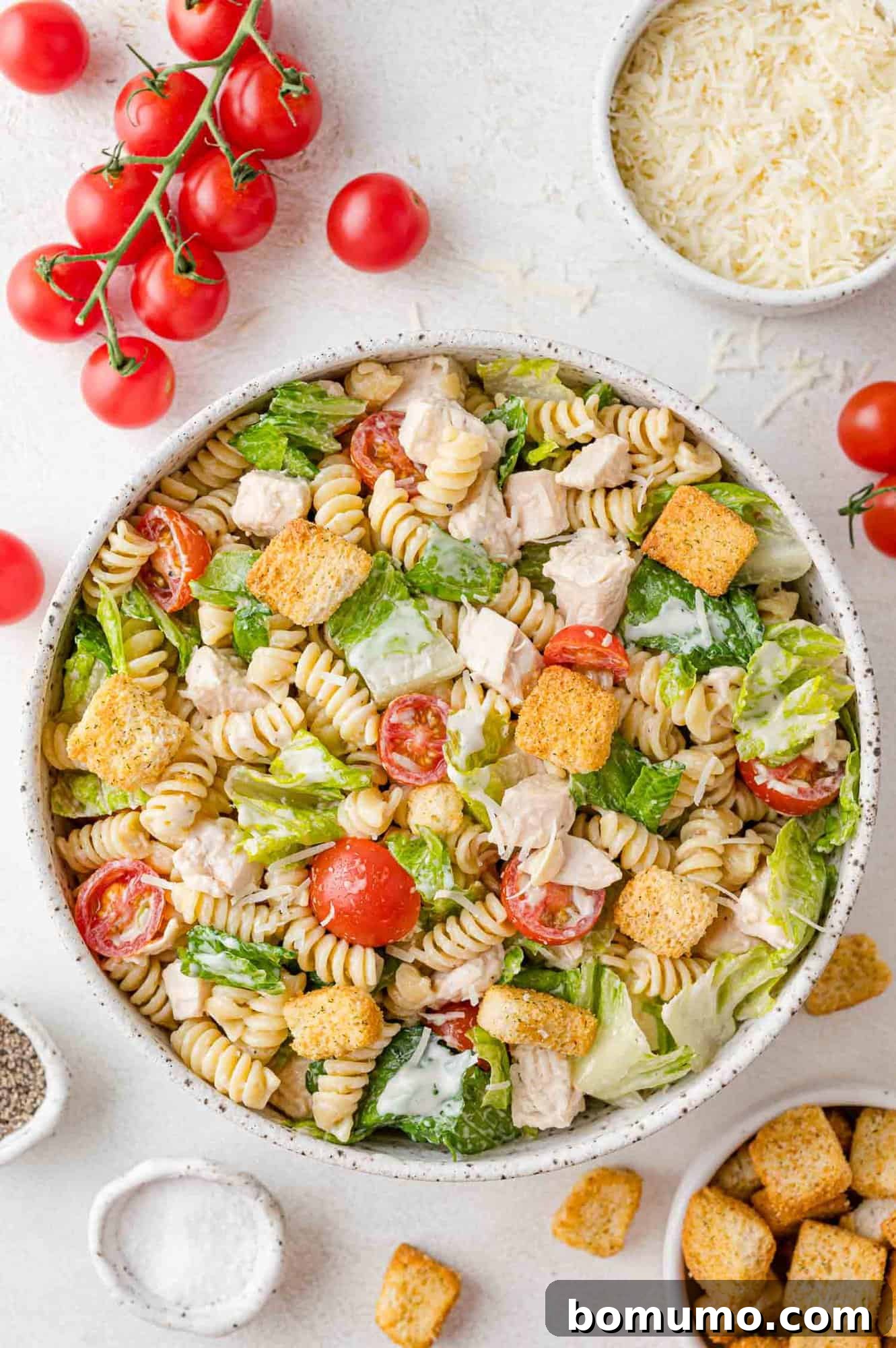 A large serving bowl filled with vibrant Chicken Caesar pasta salad, surrounded by fresh ingredients like romaine lettuce, cherry tomatoes, and Parmesan cheese.