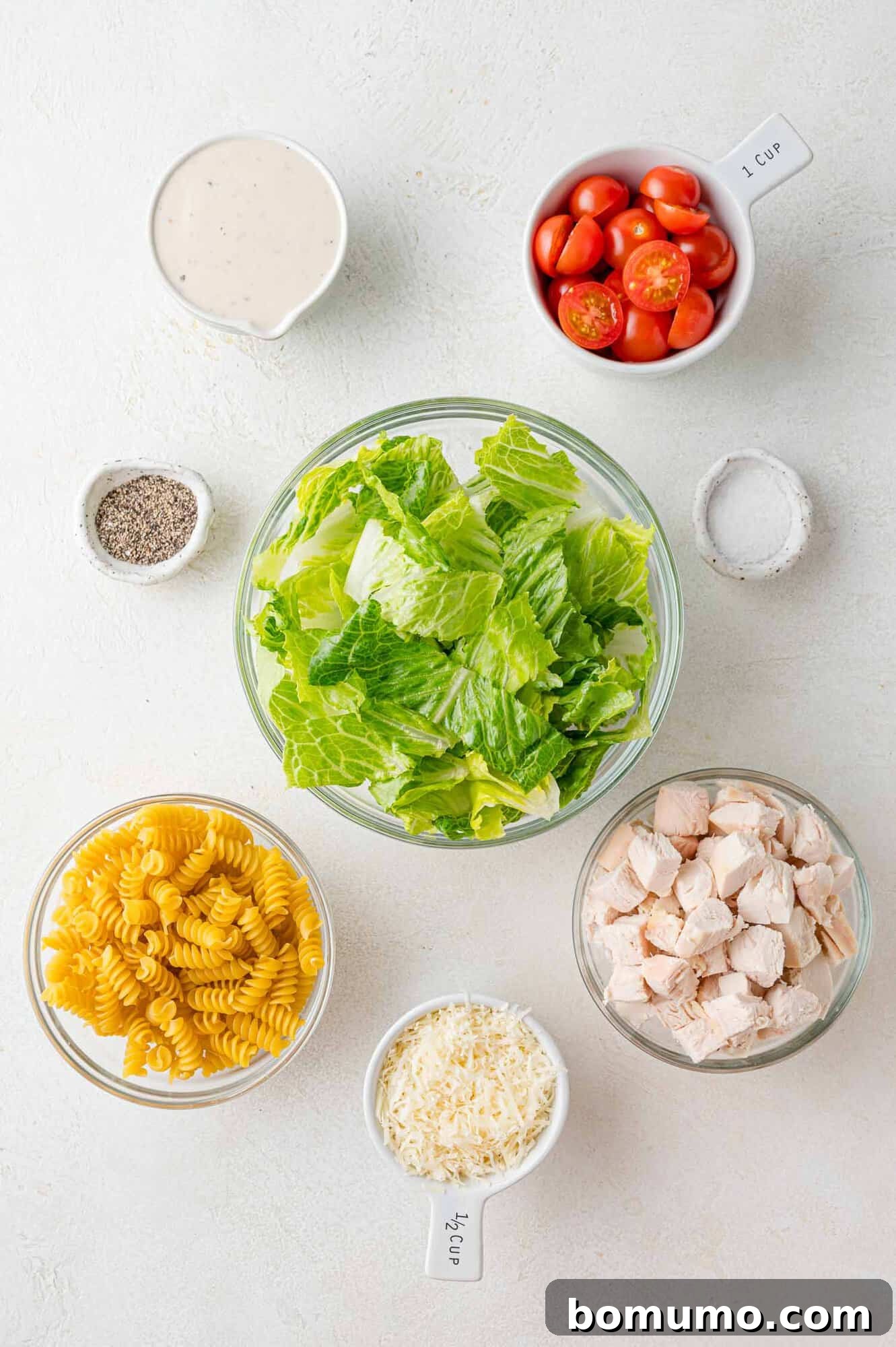 Overhead view of all the fresh ingredients meticulously laid out for the Chicken Caesar pasta salad recipe, including uncooked rotini, crisp romaine, cherry tomatoes, and Parmesan.