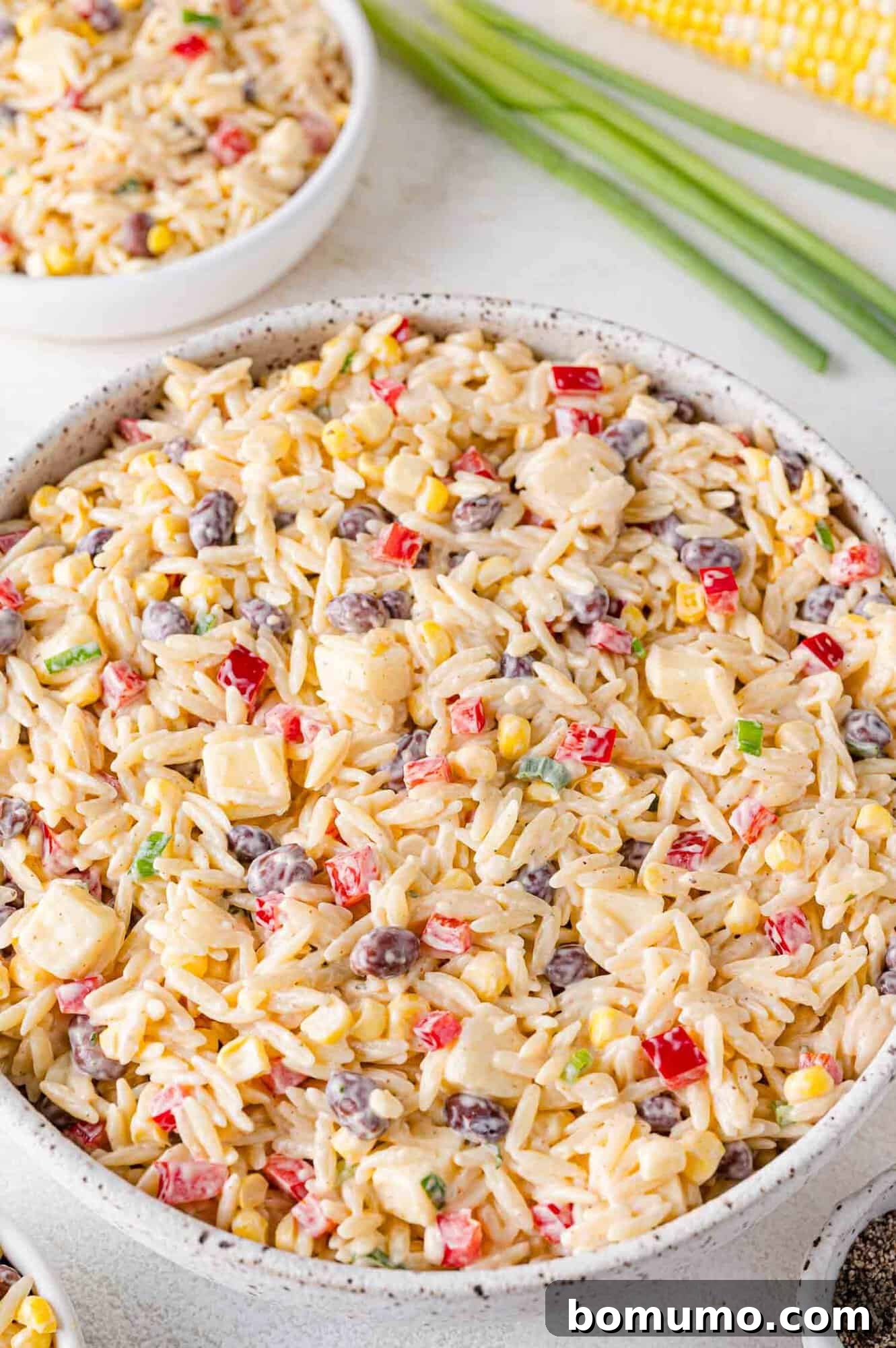 A large bowl filled with creamy southwestern orzo salad, featuring visible black beans, corn, red bell peppers, and cheddar cheese cubes.