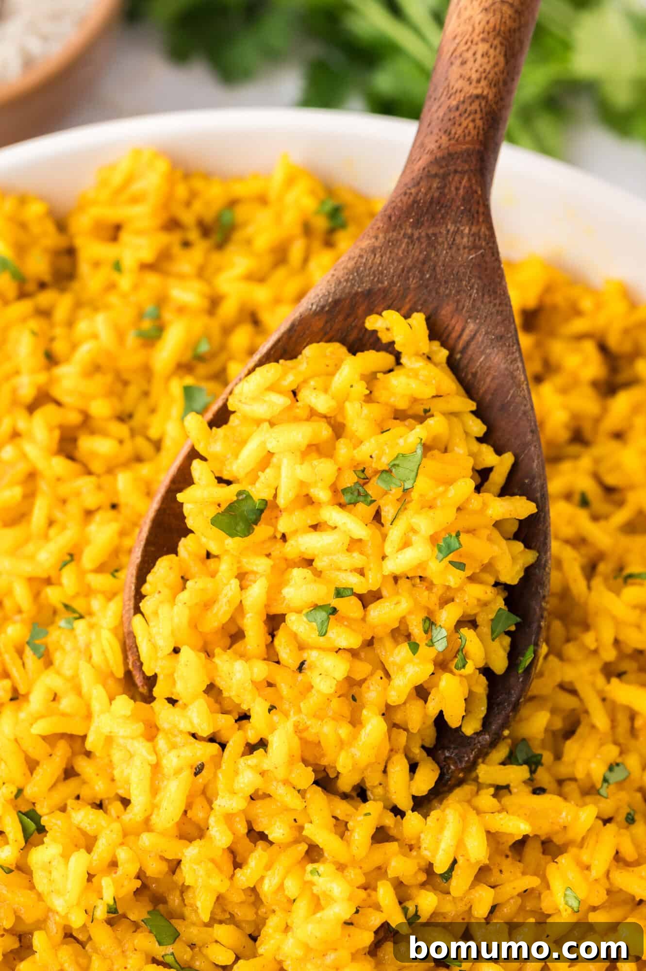 A wooden spoon scooping seasoned yellow rice garnished with fresh parsley from a serving bowl, highlighting its vibrant color and fluffy texture.