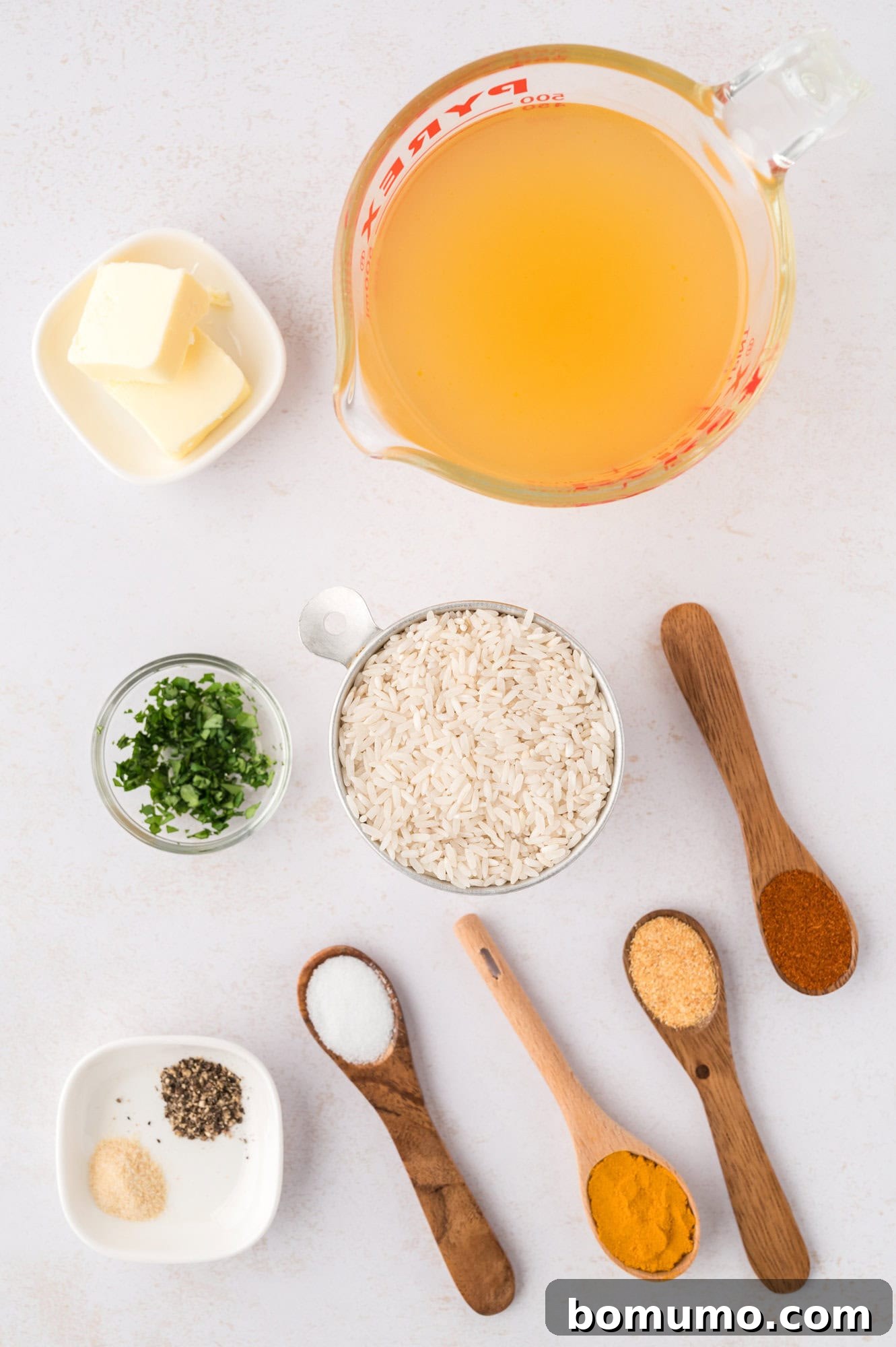 A flat lay arrangement of ingredients for yellow rice: cubes of butter, clear broth, uncooked long-grain rice, chopped green onions, black pepper, kosher salt, golden turmeric, fragrant garlic powder, and vibrant paprika, all neatly arranged on a light background.