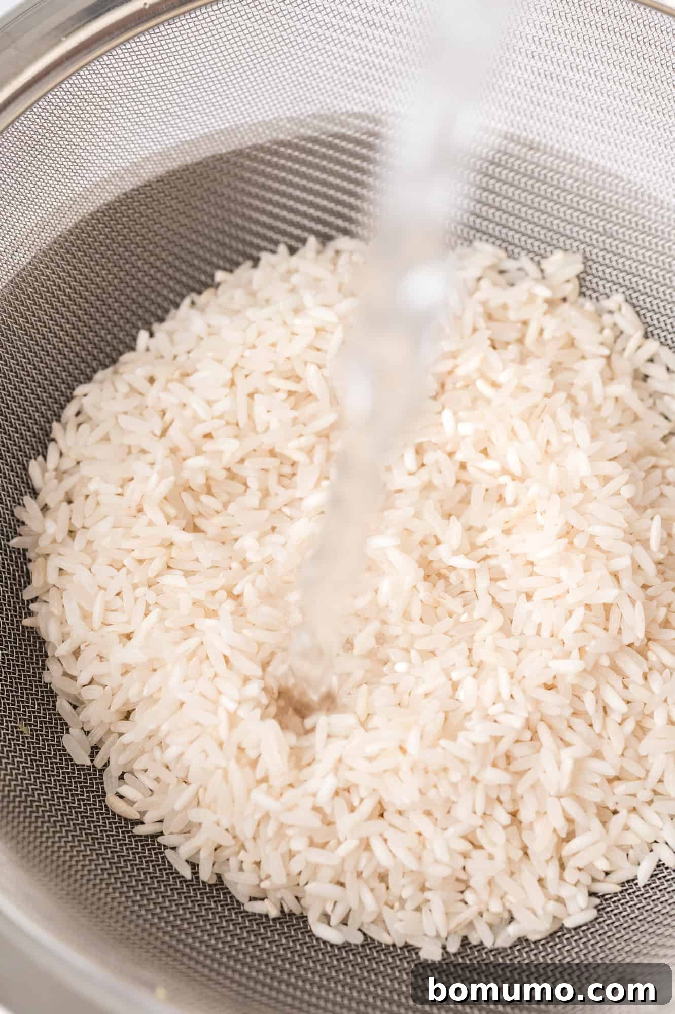 Water running over uncooked white rice in a metal strainer, illustrating the crucial rinsing step to remove excess starch.