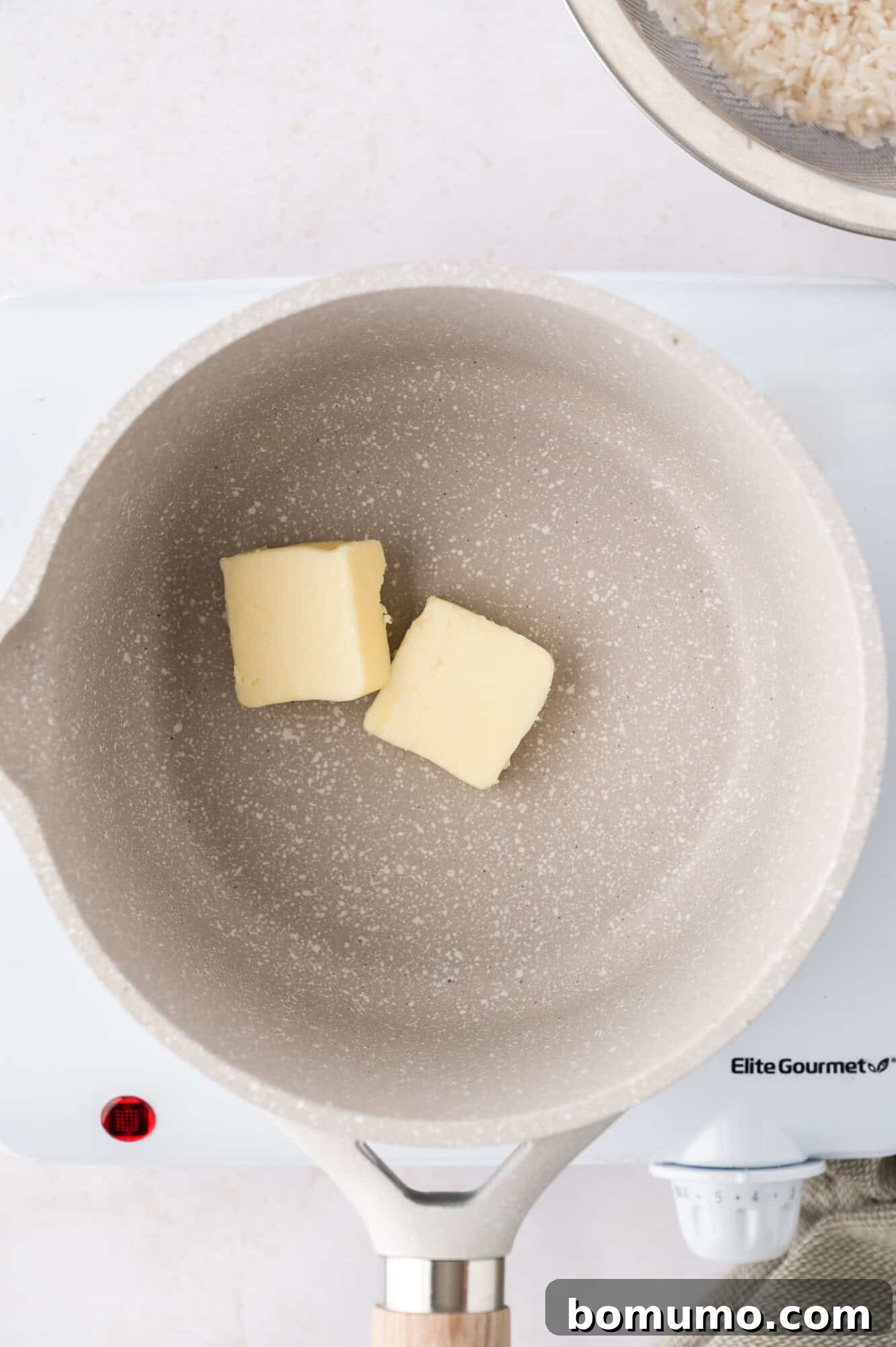 Two pats of butter melting in a light gray saucepan on a stovetop, ready for the rice to be added.
