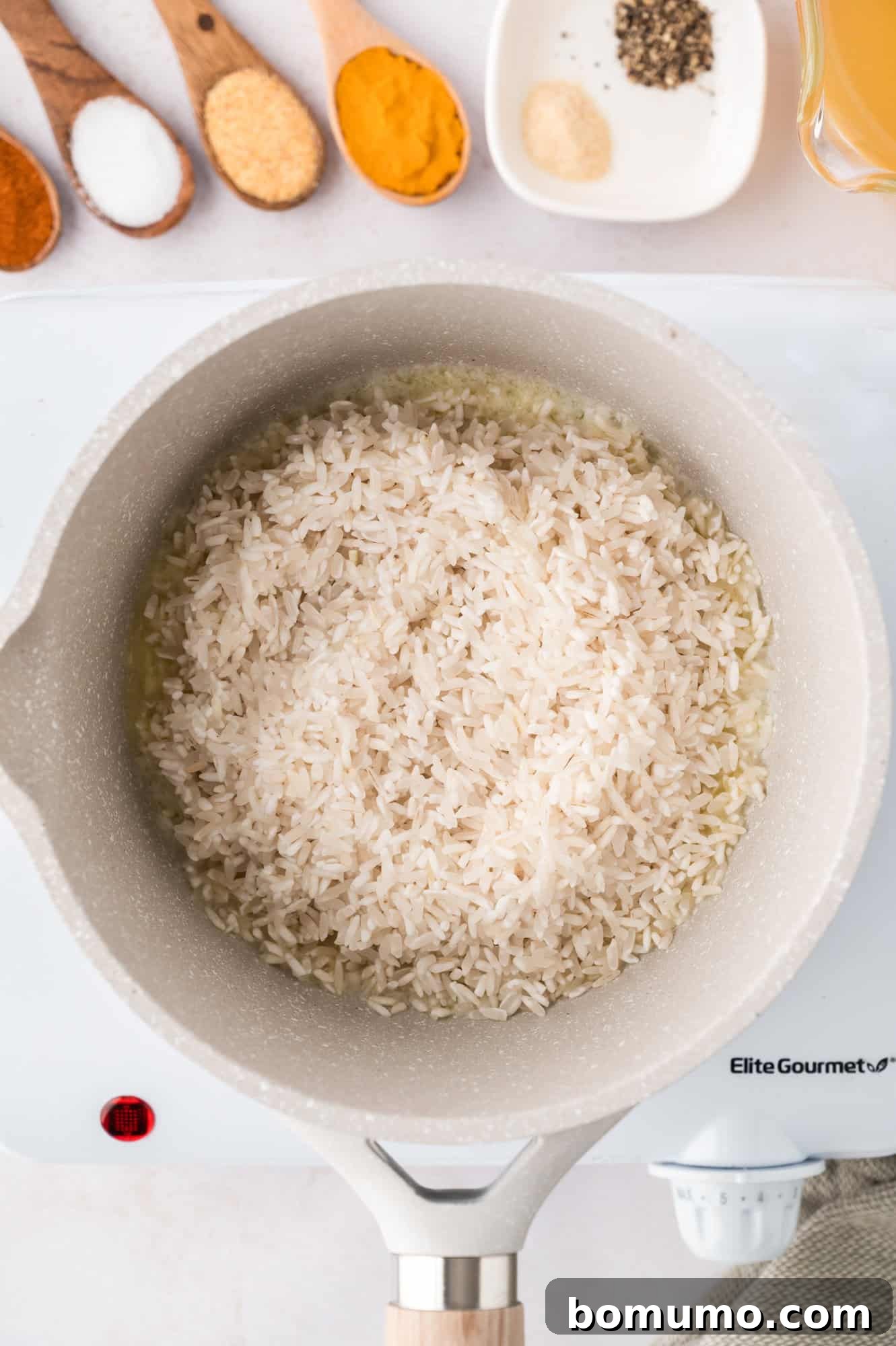 Uncooked white rice toasting in a pot on a stove, with small bowls of spices nearby, ready to be seasoned.