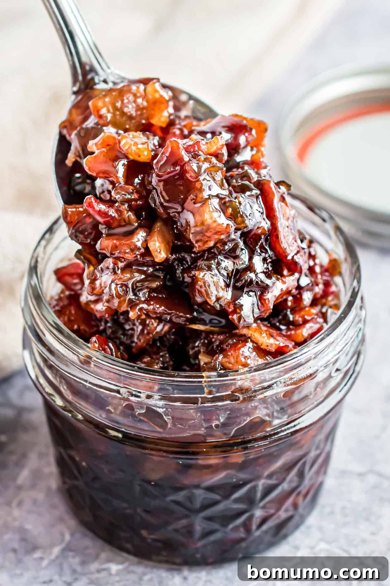 Sweet & Savory Bacon Jam 2 A small glass jar filled with rich, glossy bacon jam, with a spoon scooping out a generous portion. The jam is chunky with visible bits of bacon and caramelized onion, resting on a rustic wooden surface.