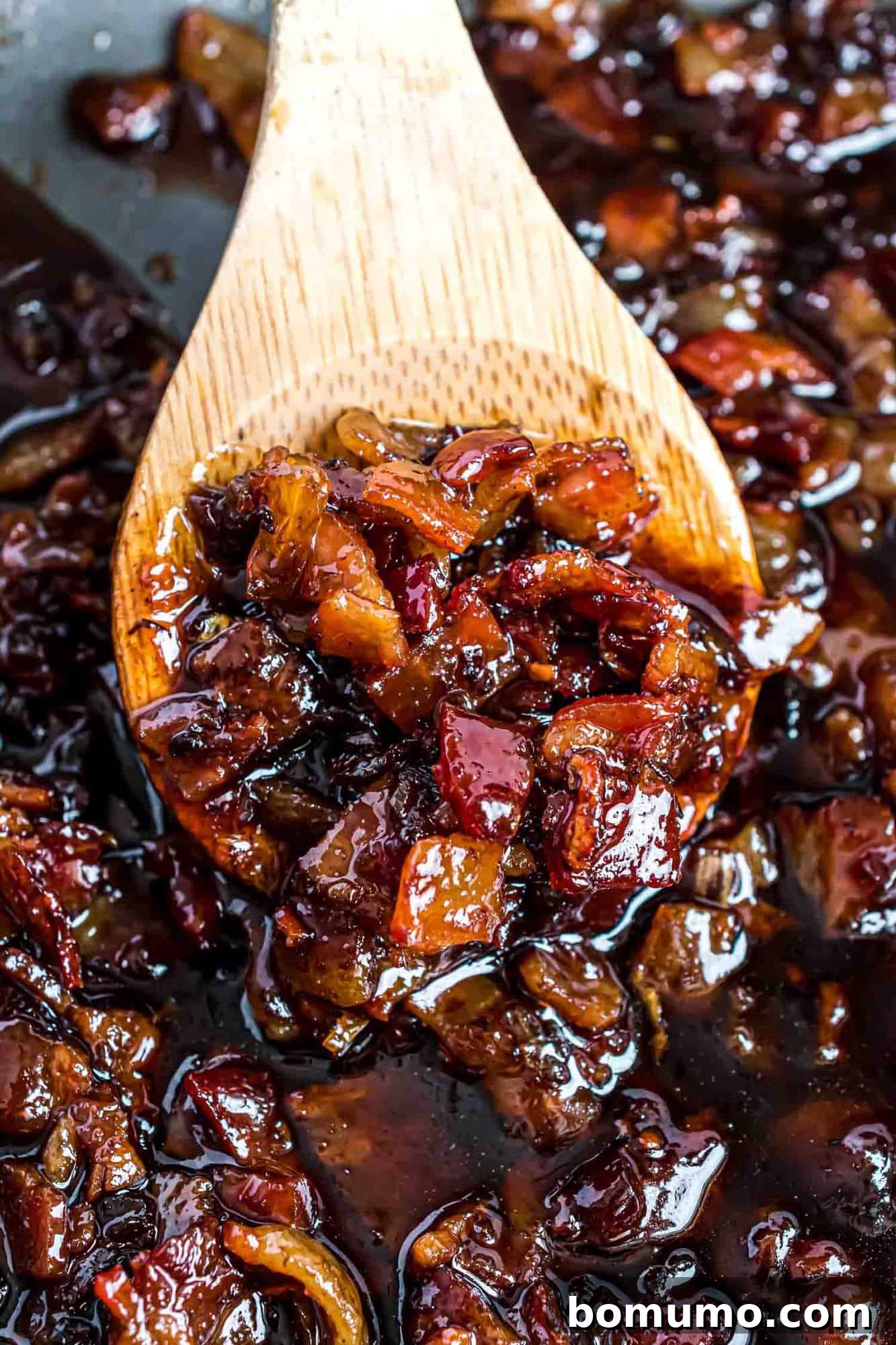 Sweet & Savory Bacon Jam 11 A close-up view of a large wooden spoon laden with a generous scoop of glossy, chunky bacon jam, showcasing its appealing texture, rich color, and delicious bits of bacon and onion.