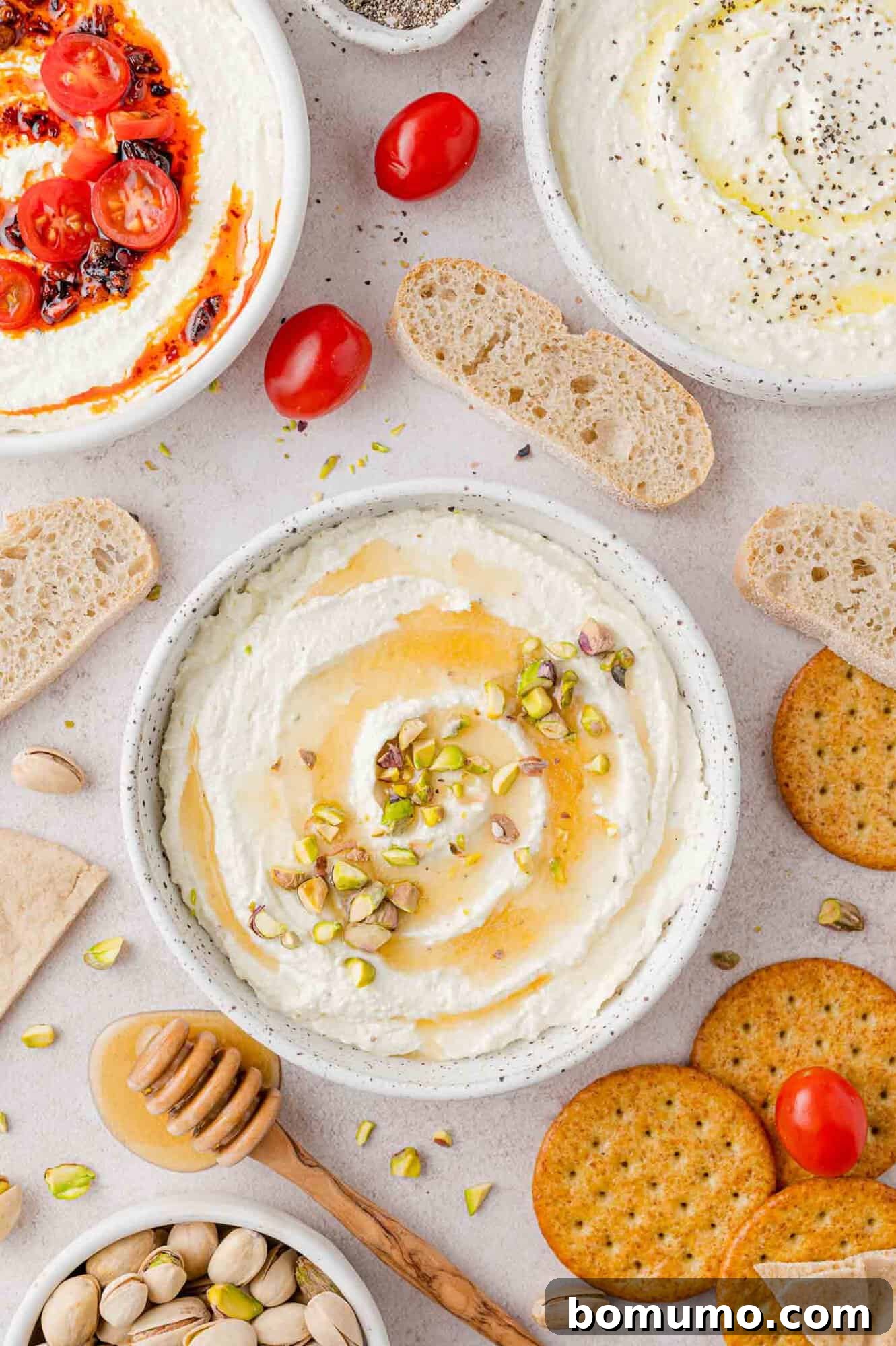 Three small white bowls of creamy whipped feta, each topped with a different garnish, surrounded by crusty bread slices and crackers, creating an inviting appetizer spread.