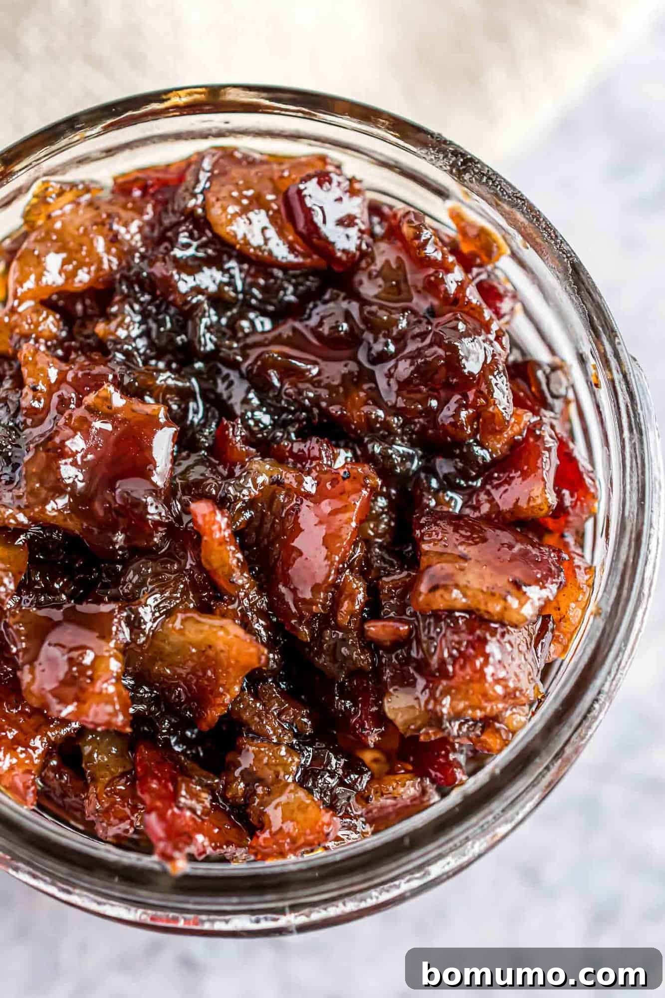 Sweet & Savory Bacon Jam 13 A small, elegant glass jar brimming with homemade bacon jam, positioned on a rustic wooden surface, highlighting its appetizing texture and rich color, ready to be enjoyed.