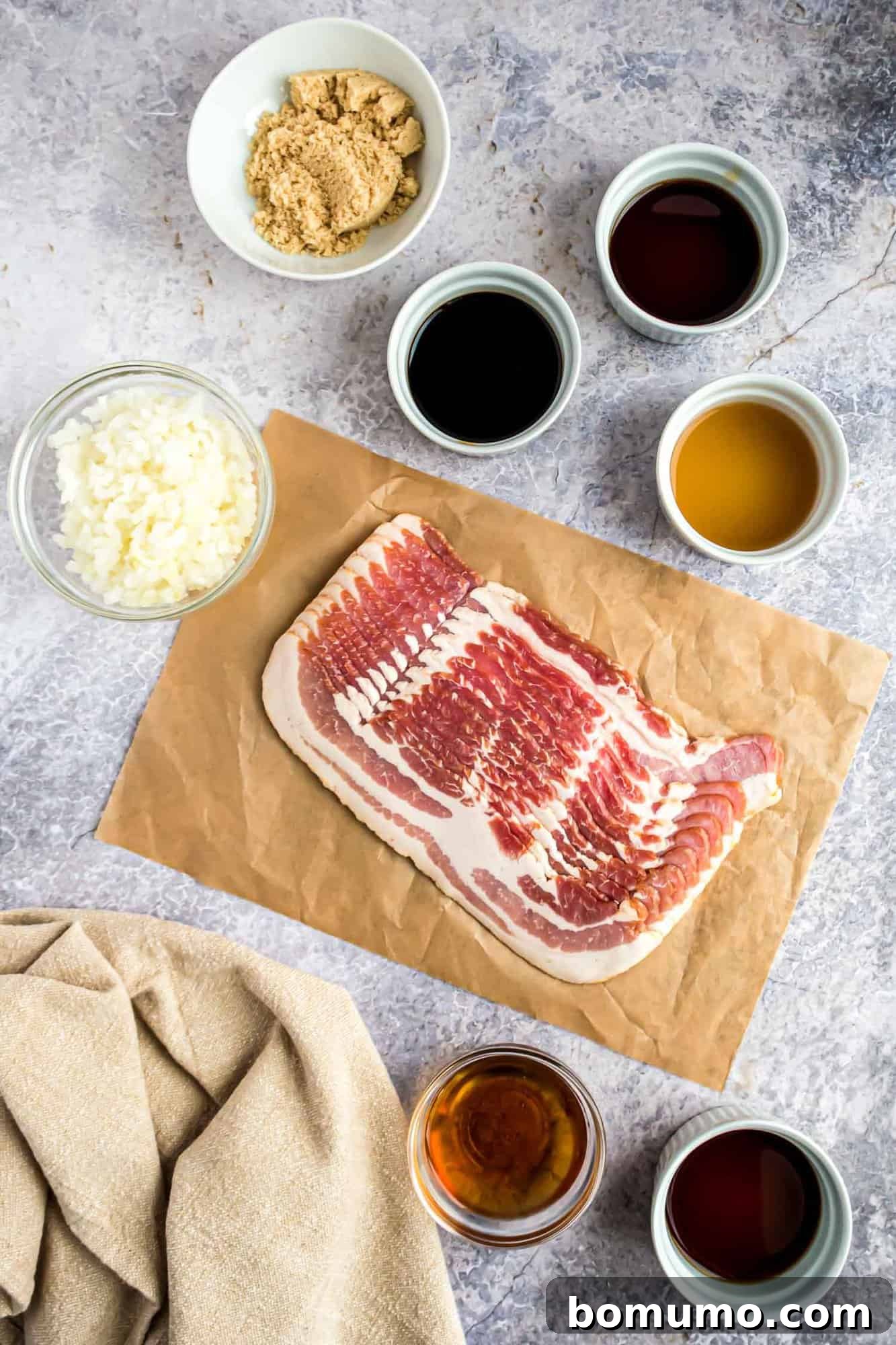 Sweet & Savory Bacon Jam 3 An overhead shot showcasing all the fresh ingredients neatly arranged on a wooden surface, including strips of raw thick-cut bacon, a whole Vidalia onion, bottles of pure maple syrup, maple bourbon, and various vinegars.