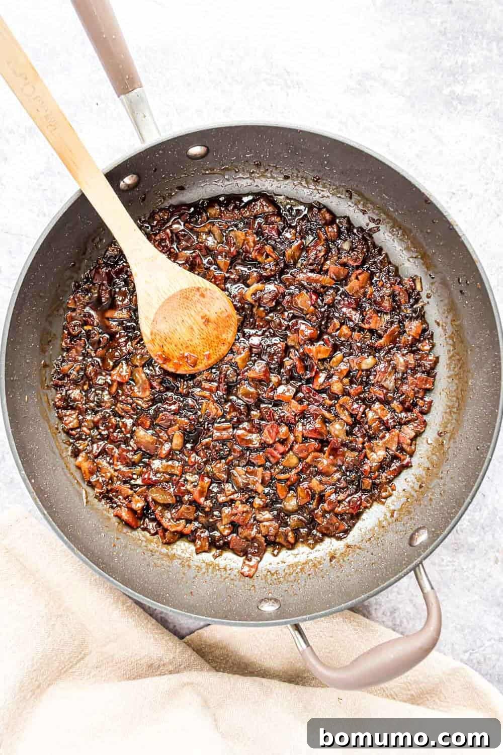 Sweet & Savory Bacon Jam 10 The finished bacon jam, rich and thick with visible bacon pieces, shown bubbling gently in the frying pan with a wooden spoon, ready to be transferred.