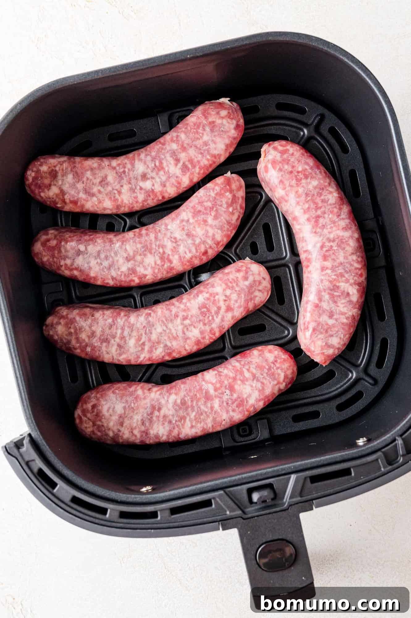 Five uncooked bratwurst sausages in an air fryer.