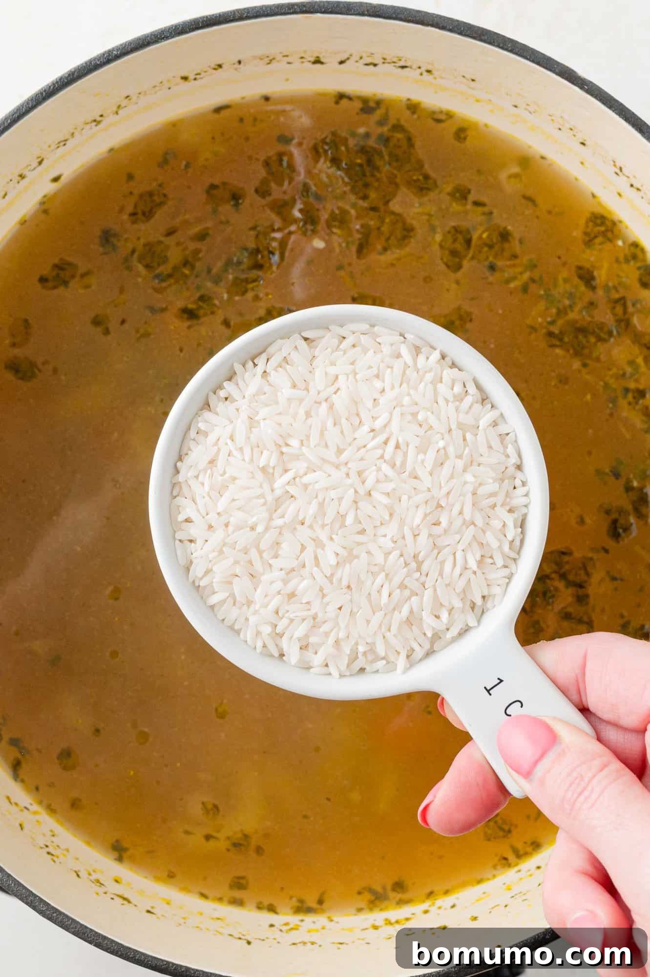 Measuring cup of rice held over pot of soup.