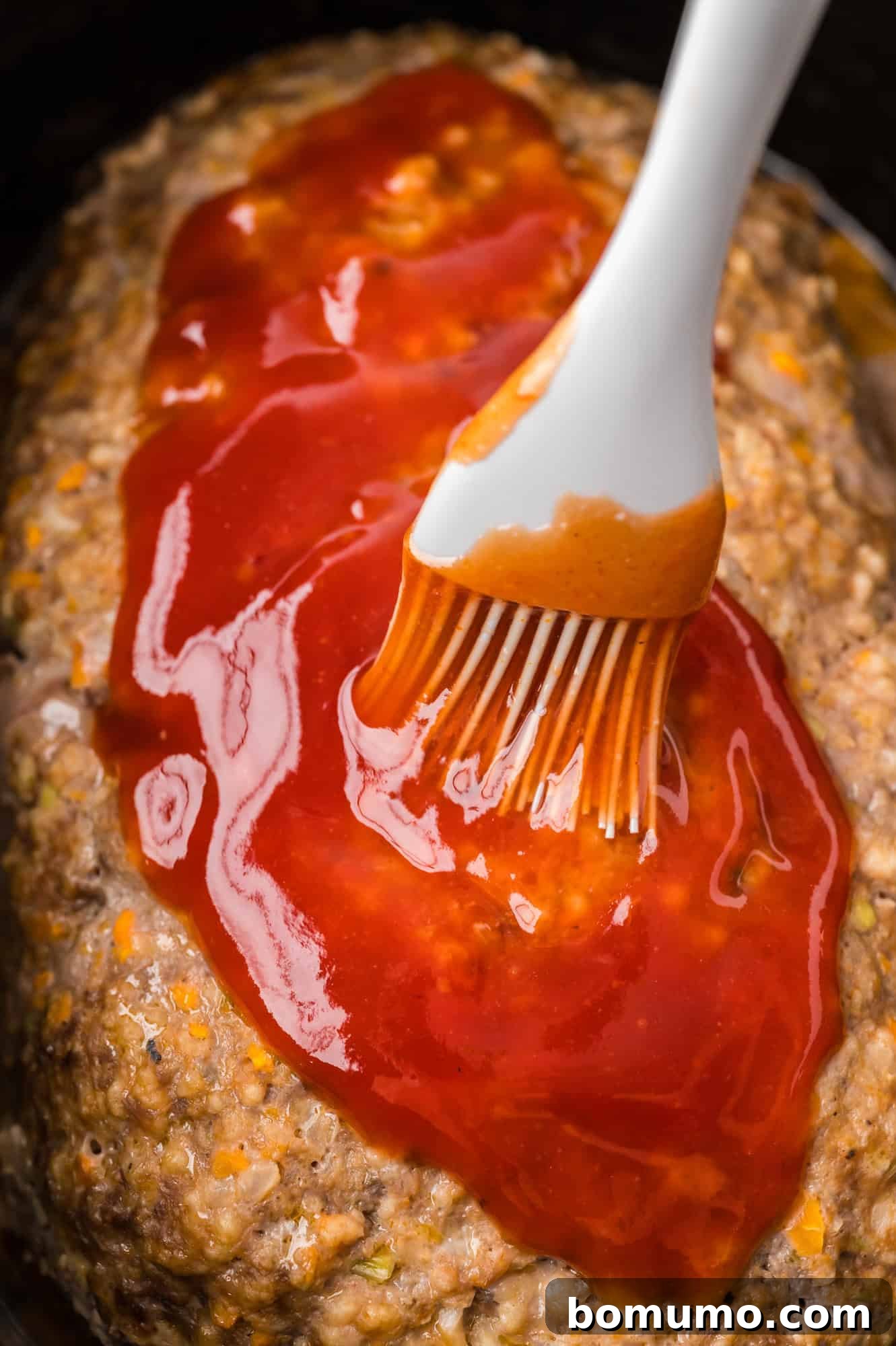 A brush applying the glistening glaze evenly over the top of the cooking meatloaf in the crockpot.