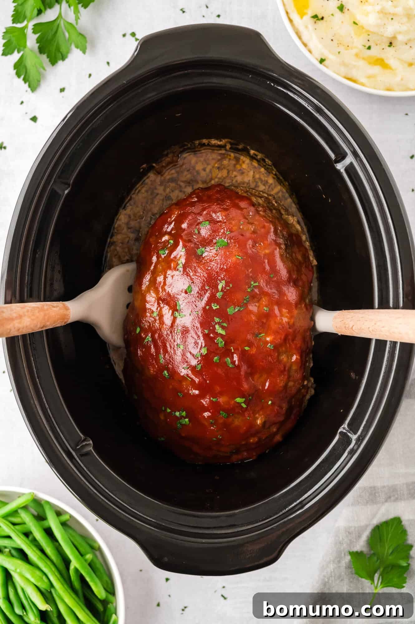Using two spatulas to carefully lift the fully cooked and glazed meatloaf out of the slow cooker.