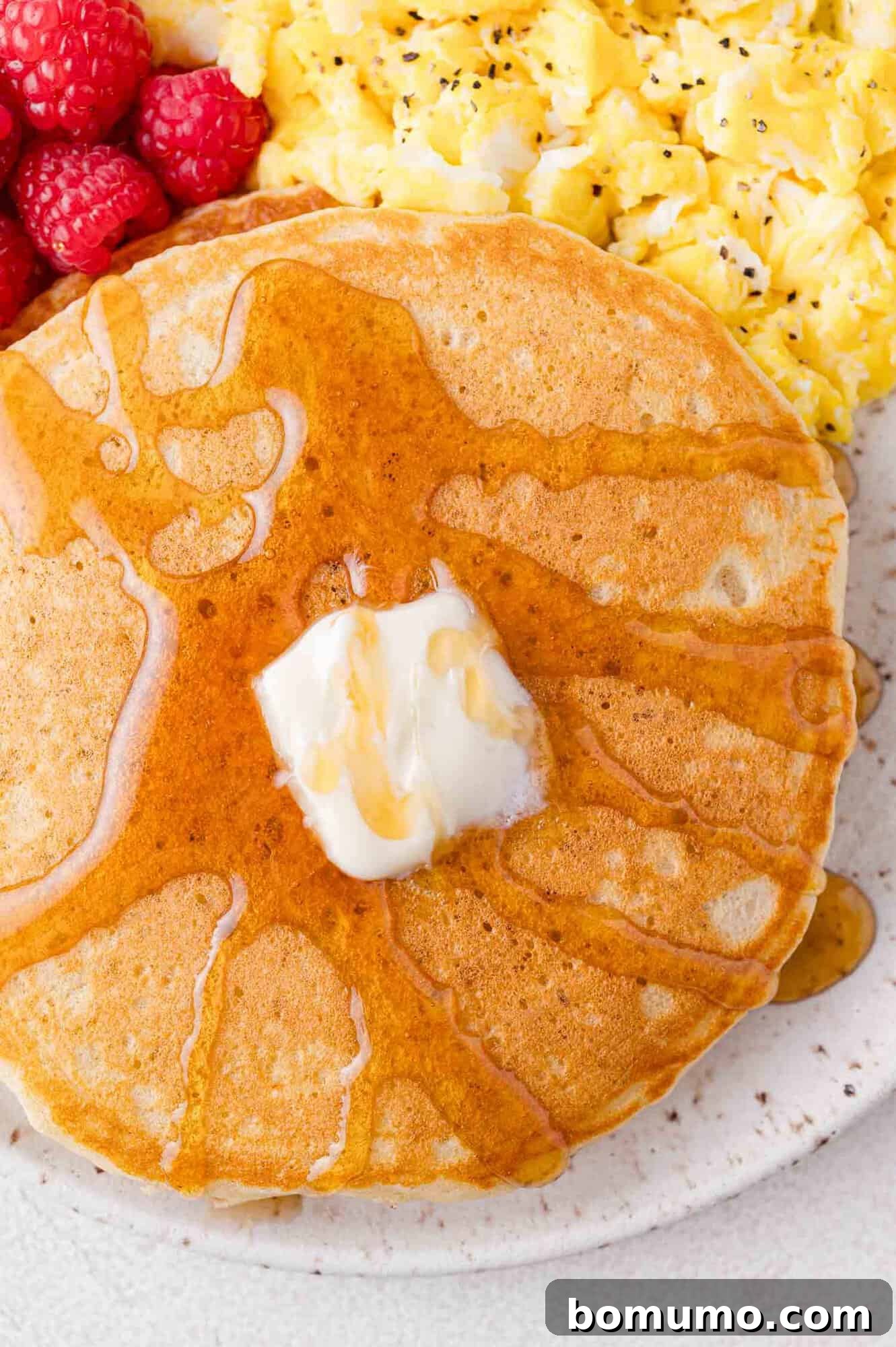 Close up of butter and syrup on pancakes, on a plate with eggs and raspberries.