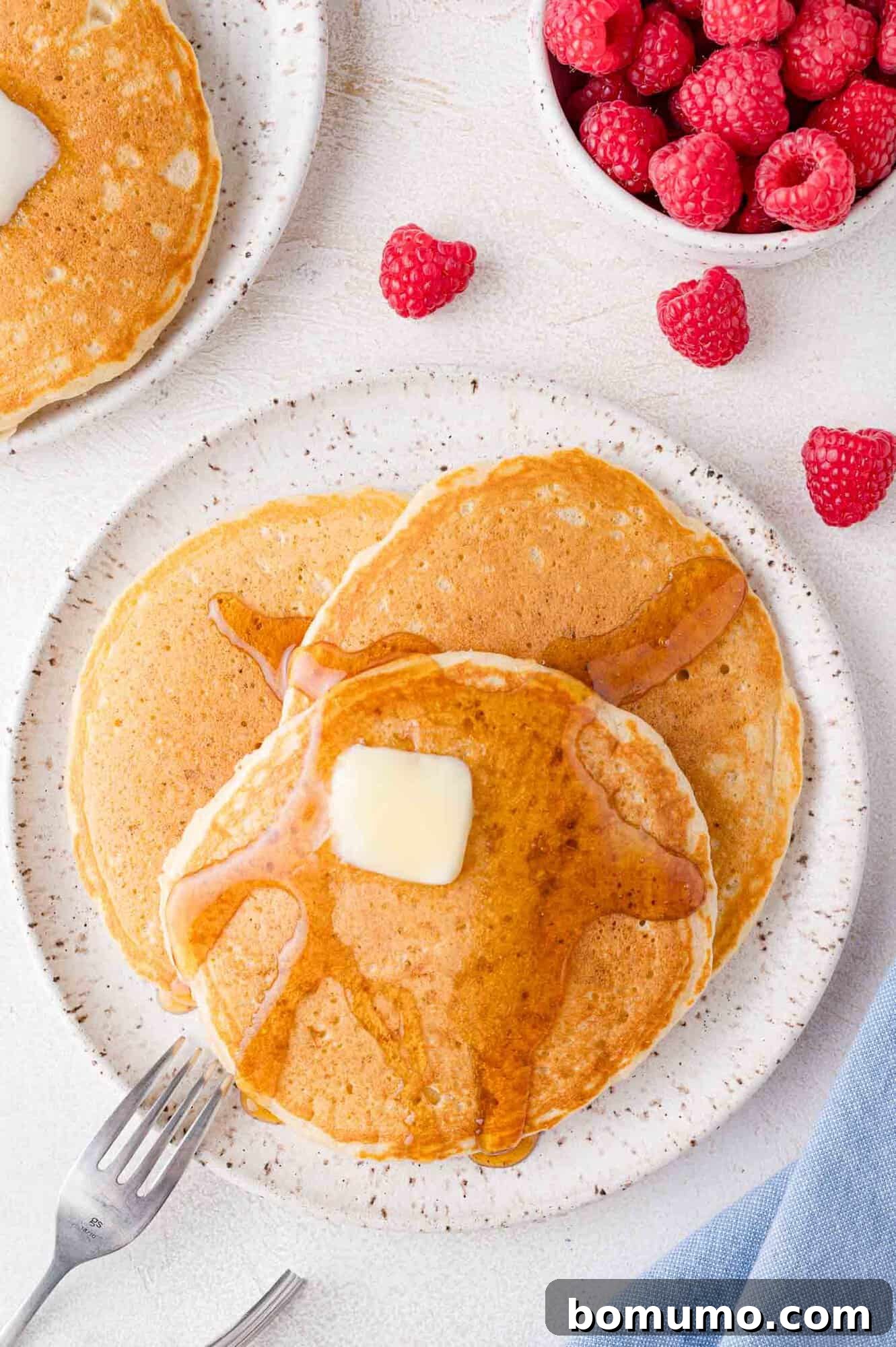 Pancakes, plated, with butter and syrup. Raspberries in the corner of the photo.