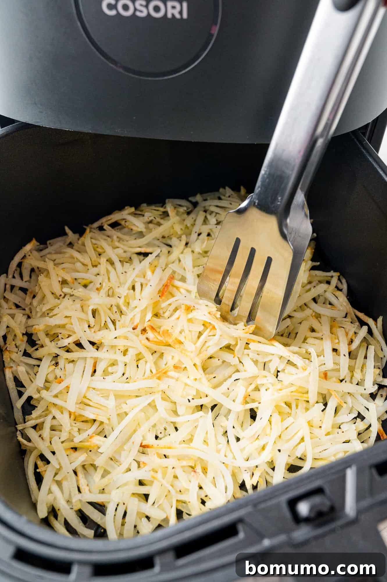 Partially cooked hash browns in an air fryer basket, being flipped with tongs to ensure even browning.