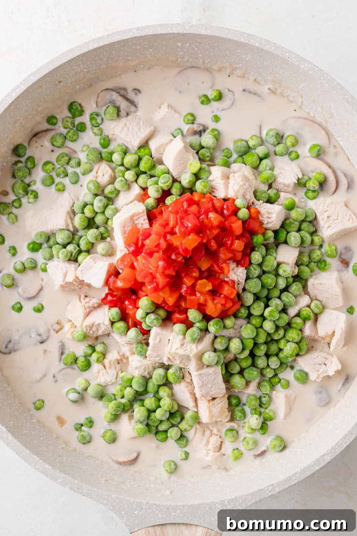 Cooked chicken, pimentos, and frozen peas freshly added to the creamy sauce in the pan.