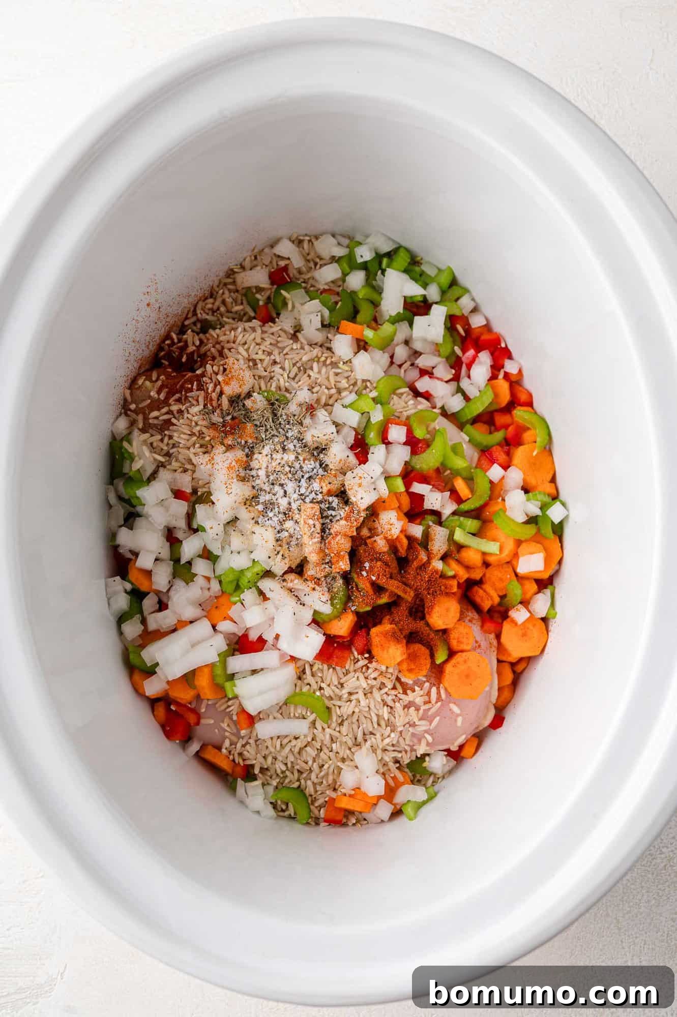 Most of the ingredients for Crockpot chicken and rice are layered in a white crockpot, before the broth is added.