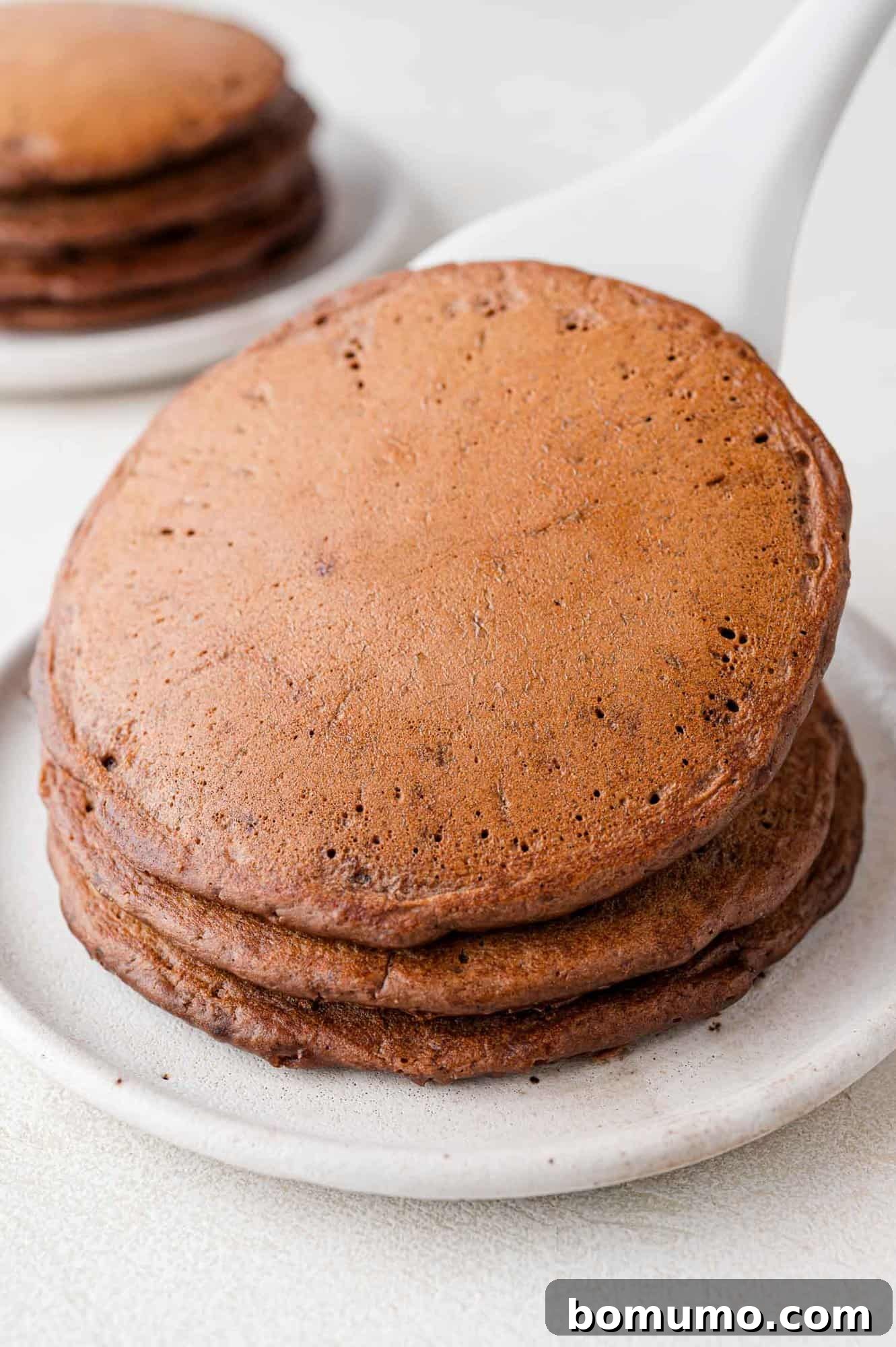 A freshly cooked chocolate pancake being added to an existing stack of two, forming a tempting tower.