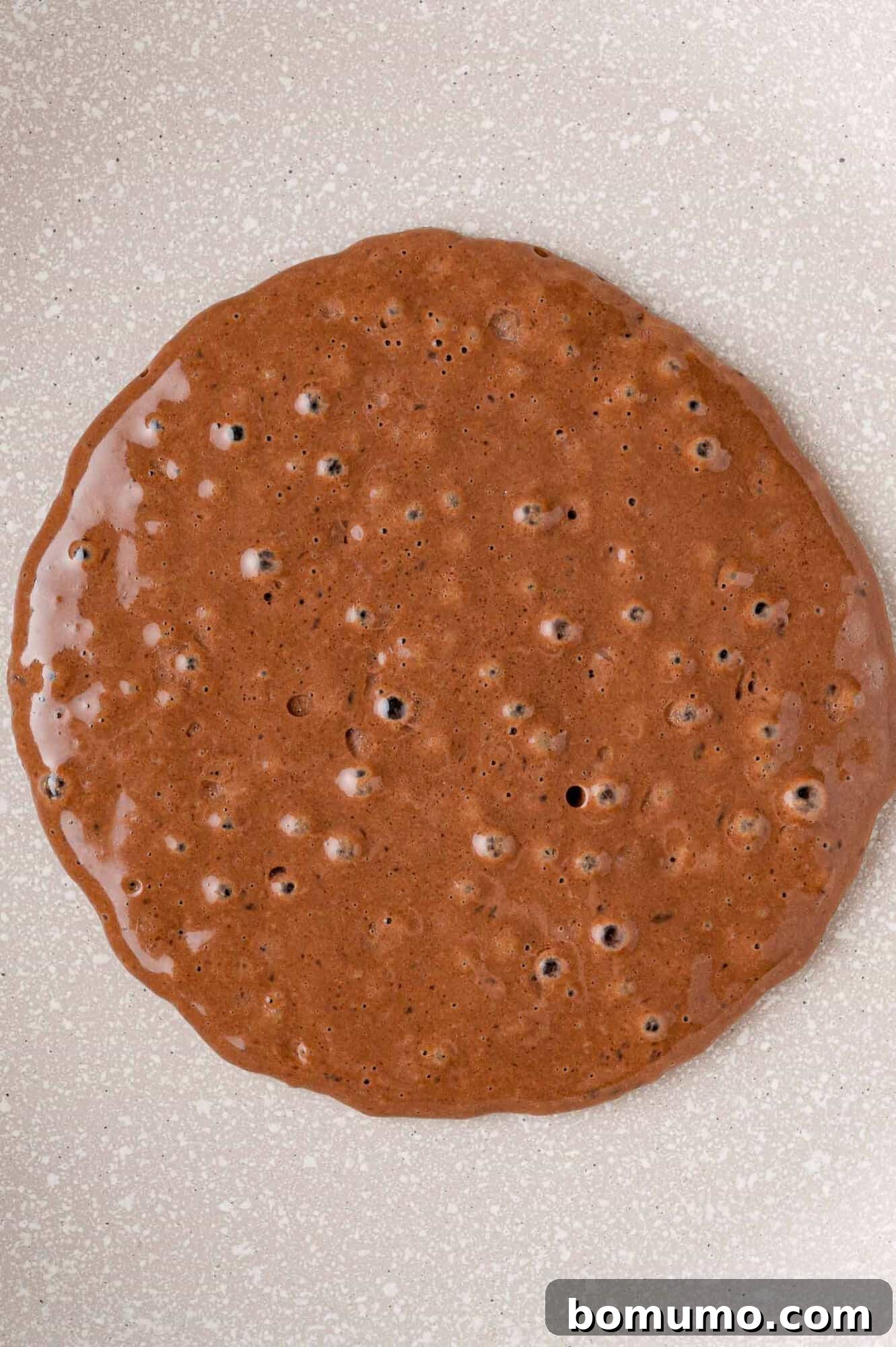 Decadent Chocolate Flapjacks 10 A chocolate pancake cooking on a griddle, with small bubbles just beginning to form on its surface.