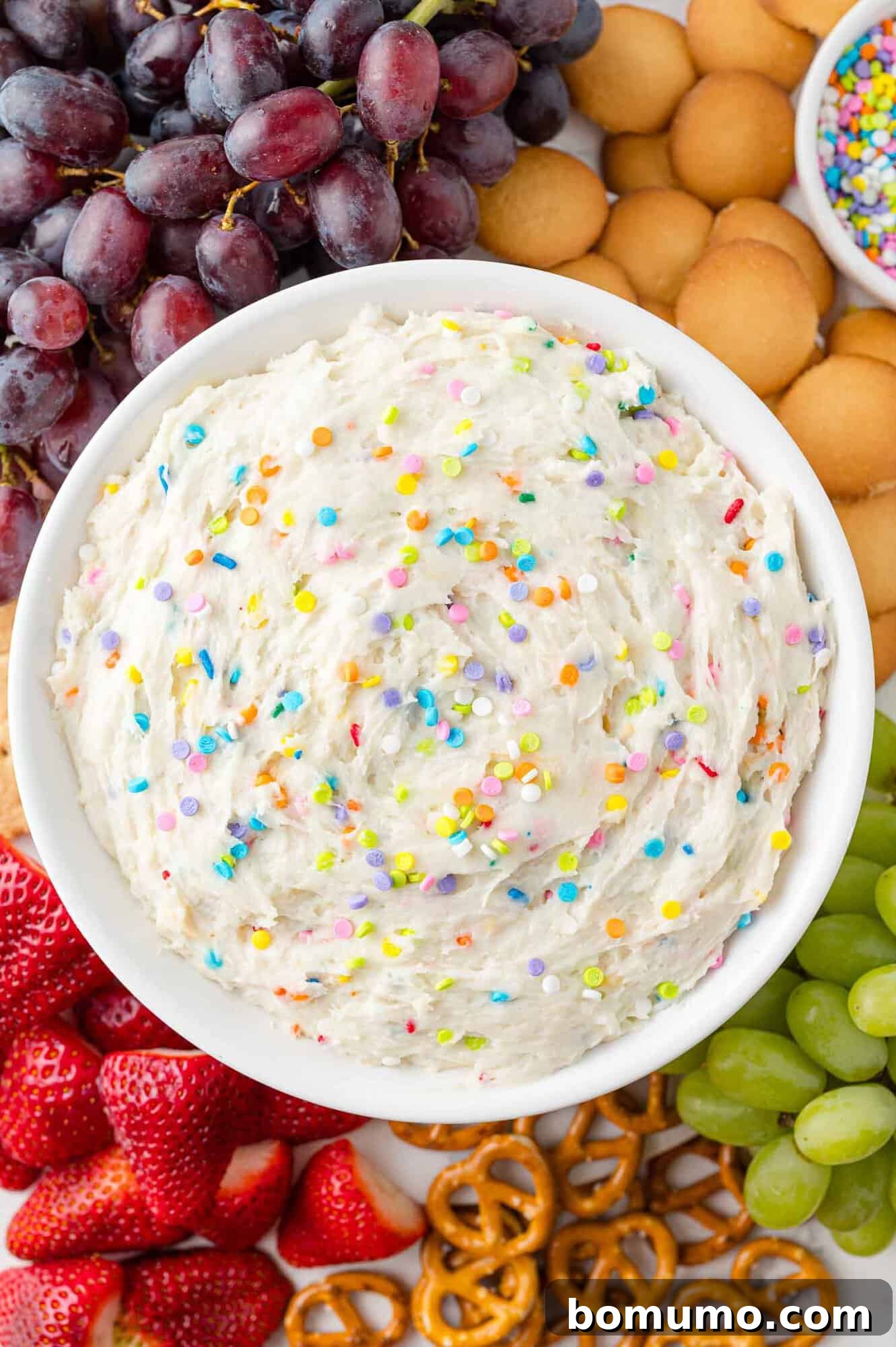 A beautifully arranged bowl of vibrant funfetti dip, generously topped with sprinkles and surrounded by a tempting assortment of dippers including crisp vanilla wafers, juicy strawberries, fresh green grapes, and crunchy pretzels. This dessert dip is perfect for any celebration.