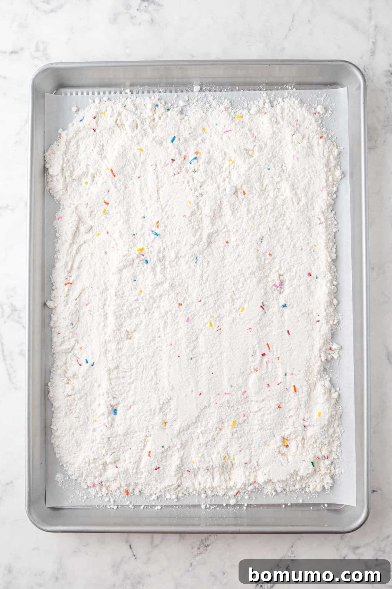 A wide, rimmed baking tray generously filled with a thin, even layer of Funfetti cake mix, resting on a polished marble surface. The cake mix is ready for its crucial heat-treating step in the oven, ensuring food safety and enhanced flavor for the dip.