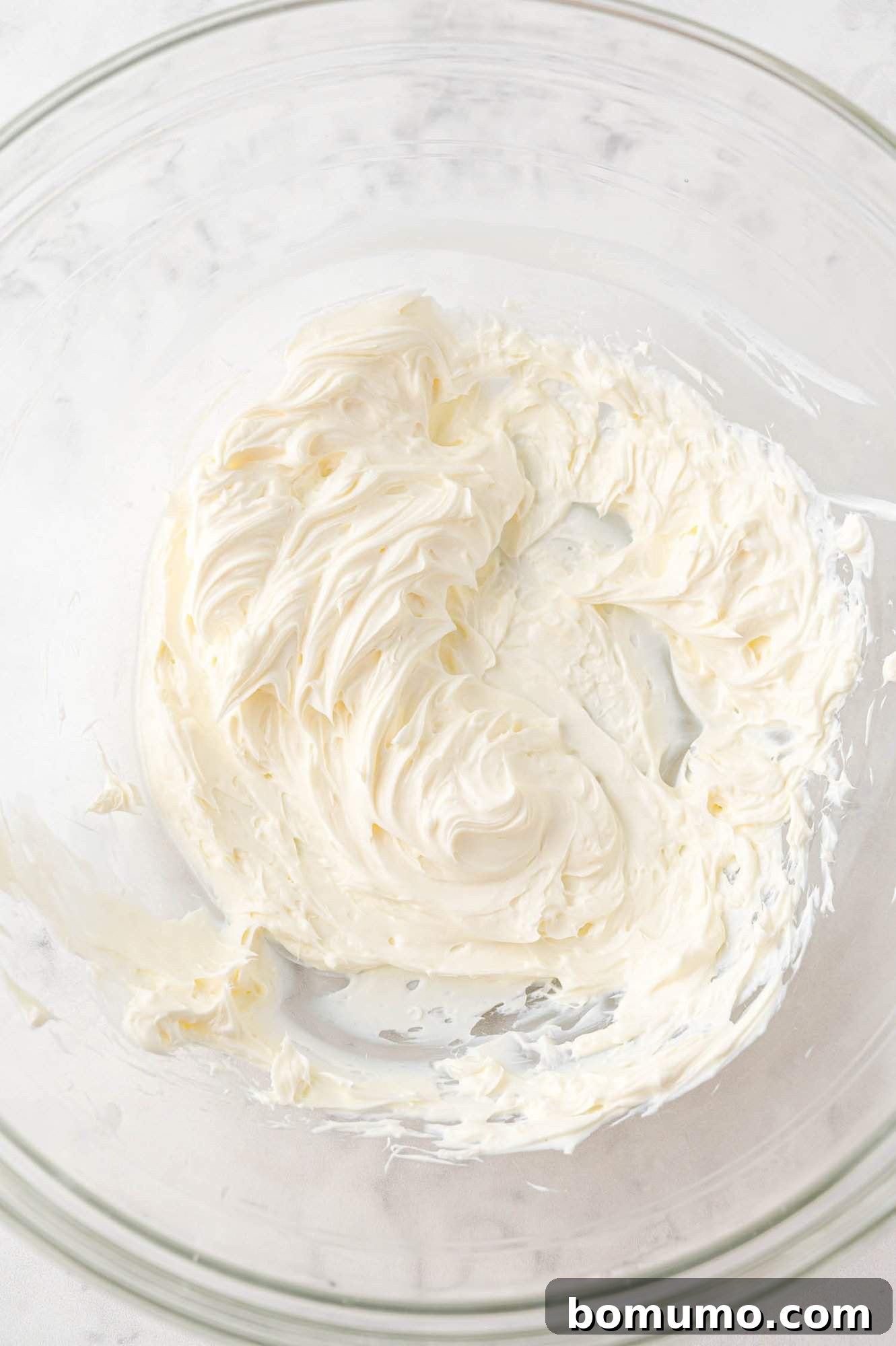 A pristine glass mixing bowl holds a generous portion of freshly whipped cream cheese, showcasing its perfectly smooth, airy, and fluffy texture. The softened cream cheese has been expertly beaten, forming a lump-free foundation for the vibrant Funfetti Dip.