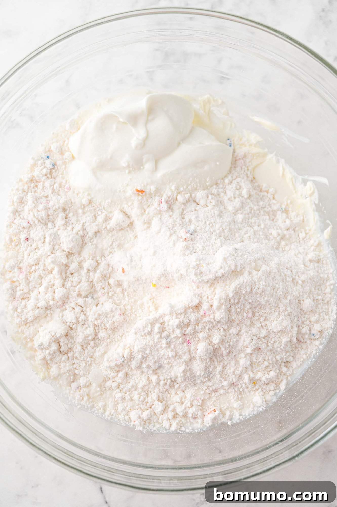 A vibrant mixing bowl is filled with the foundational ingredients for Funfetti Dip, including the fluffy white cake mix, softened cream cheese, and creamy Greek yogurt. These components are poised on a sleek marble surface, ready to be blended into a smooth, irresistible dessert.