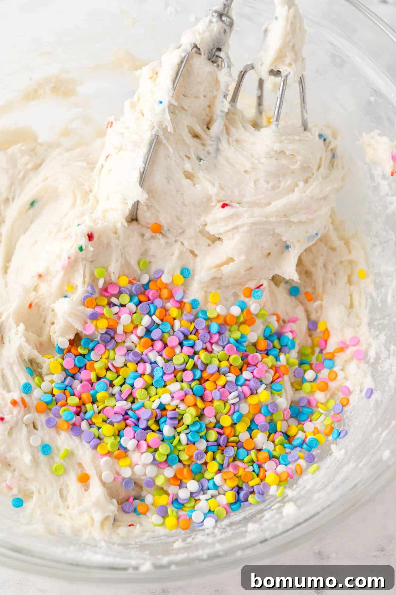 A close-up shot of a mixing bowl containing a creamy, light-colored cake batter dip, generously studded with a colorful array of sprinkles, captured mid-preparation with an electric mixer still in the bowl. This image perfectly conveys the festive and appealing nature of the Funfetti Dip.