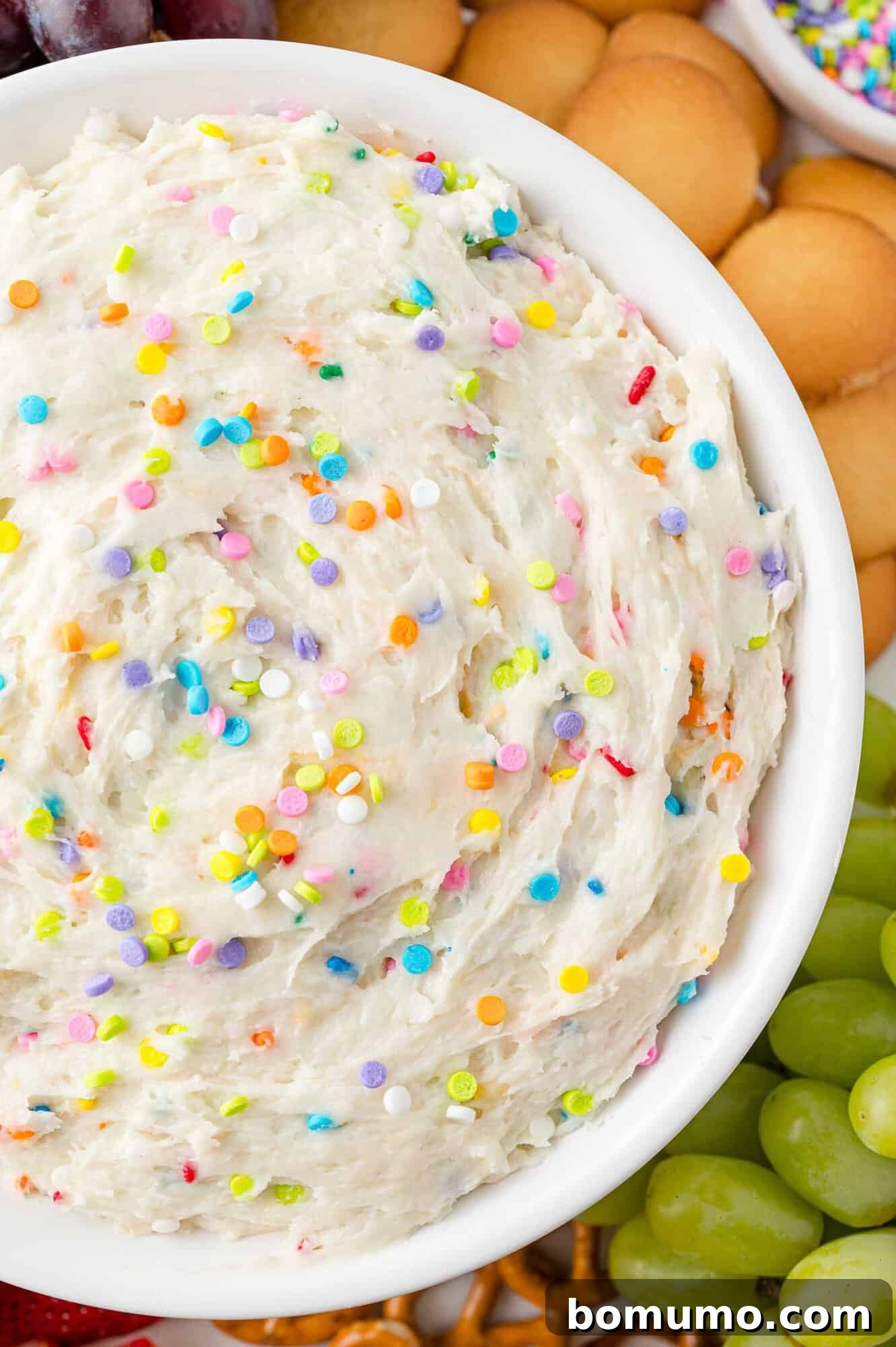 A festive bowl of funfetti dip, liberally adorned with colorful sprinkles, positioned center-frame. It's elegantly surrounded by a tempting array of dippers including classic vanilla wafers, fresh green grapes, and crunchy pretzel twists, creating a perfect party spread.
