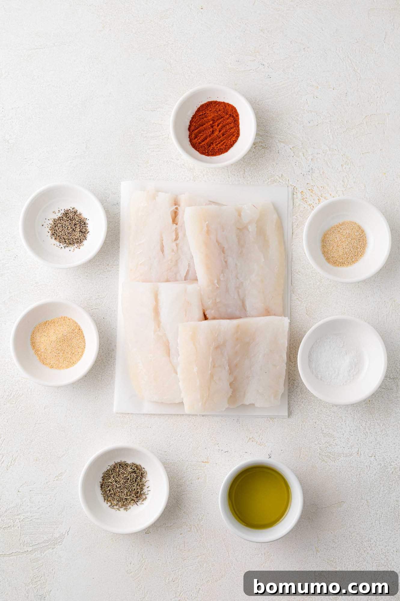 Raw cod fillets on a plate surrounded by small bowls of spices, herbs, and olive oil on a white surface.