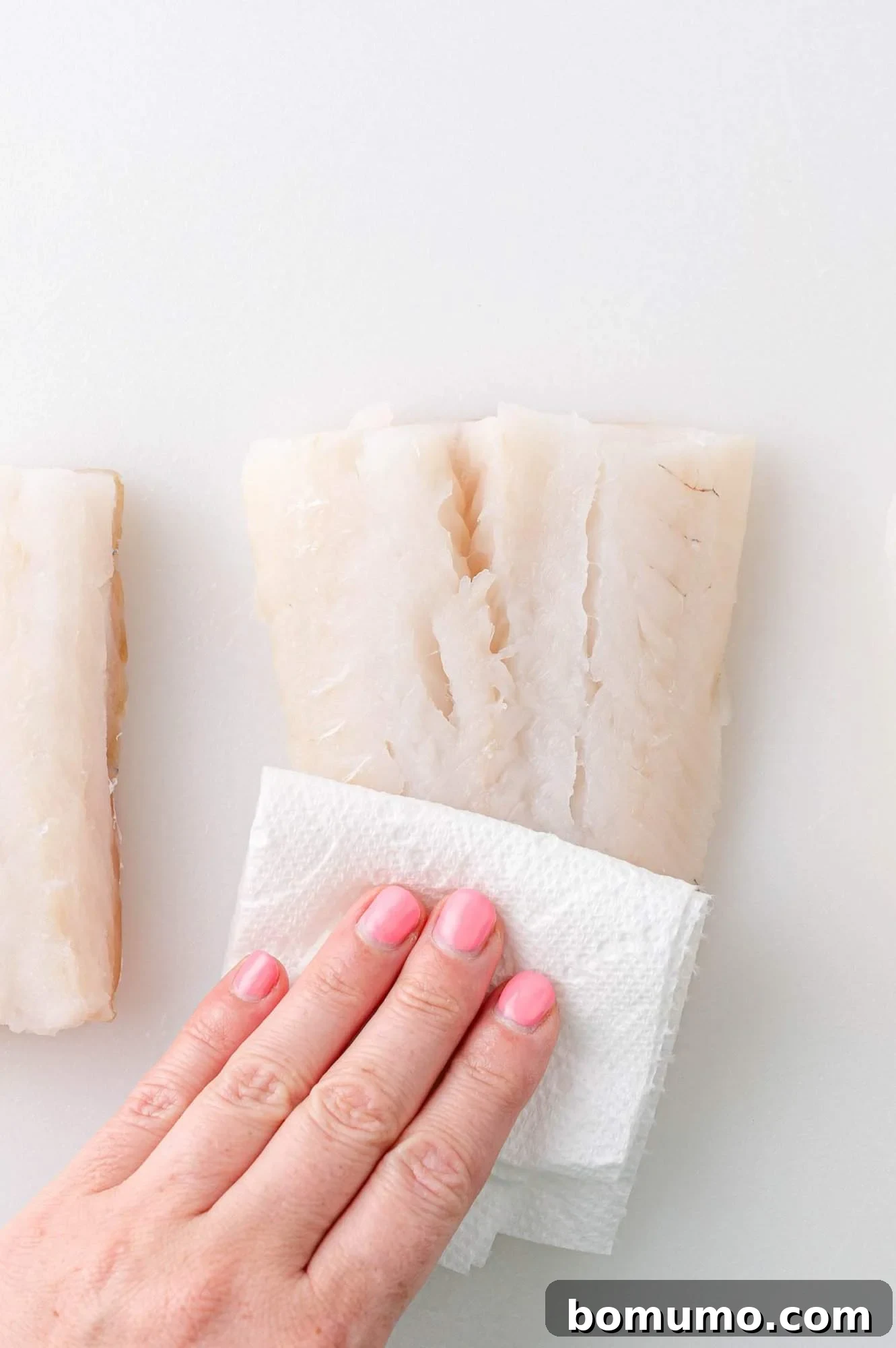 A hand pats dry a piece of raw fish with a paper towel on a white surface.