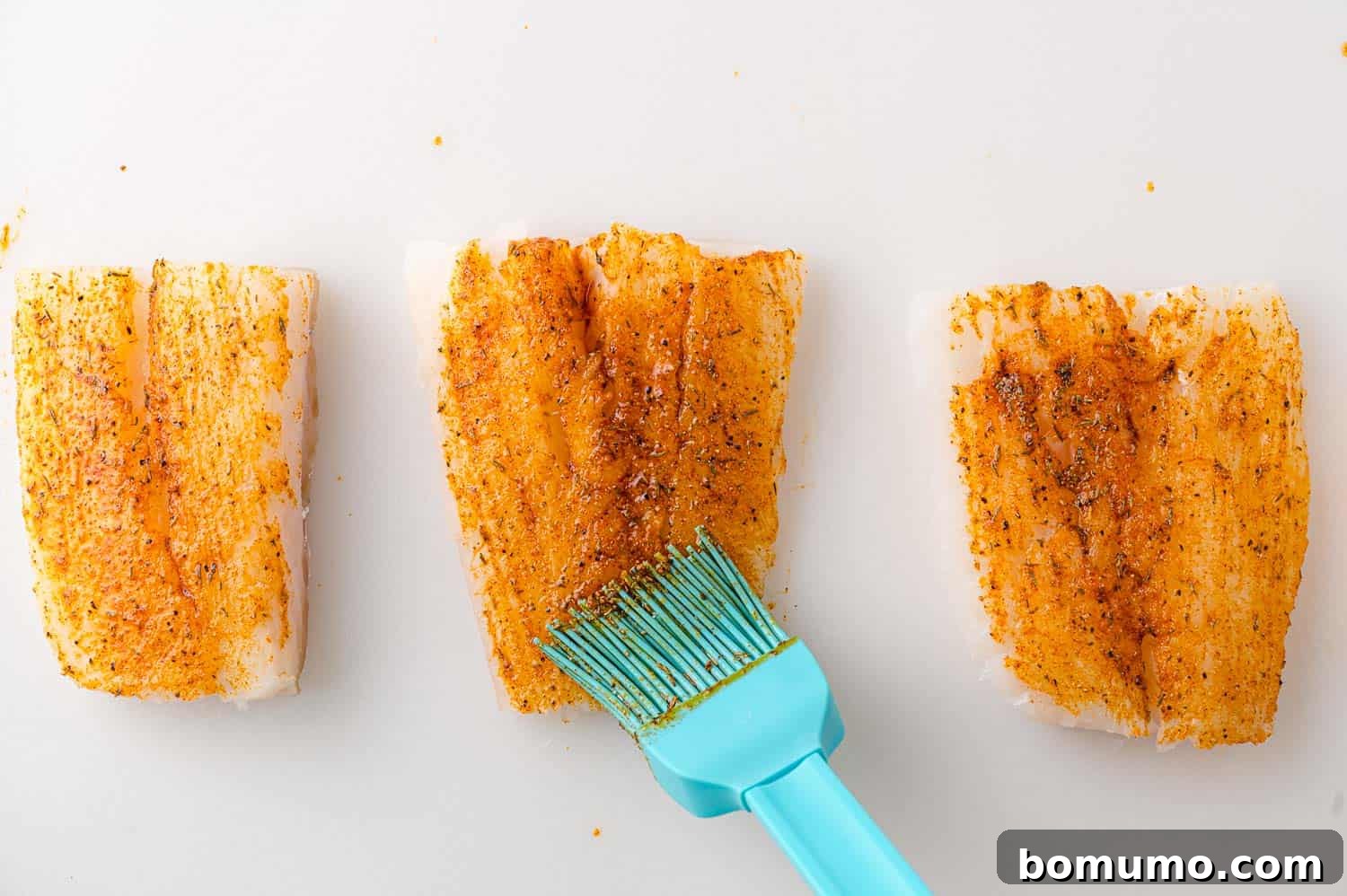 Cod fillets on a white surface being brushed with spices using a blue silicone brush.