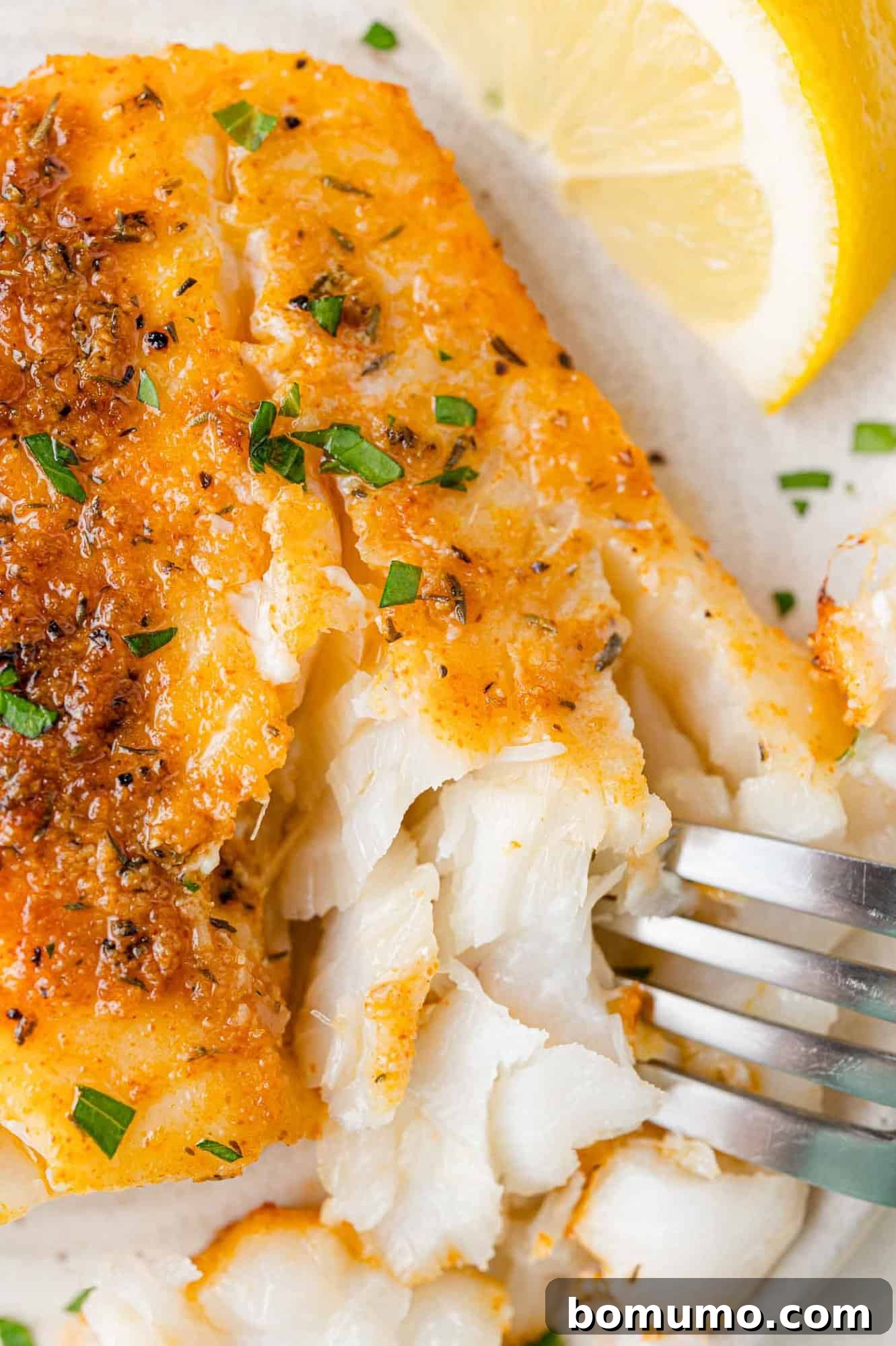 Close-up of a fork flaking a cod fillet garnished with herbs, next to a lemon wedge.