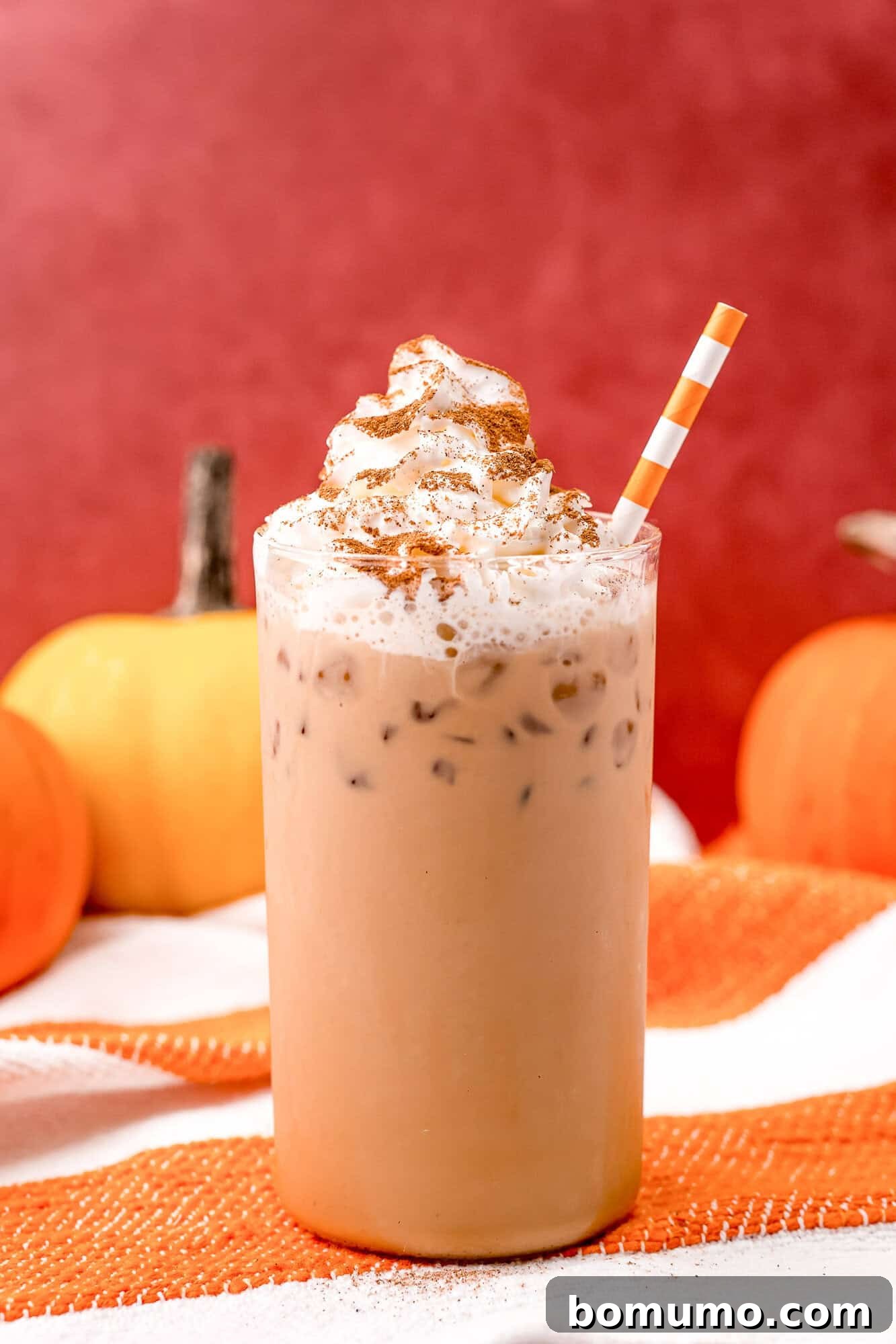 Refreshing Iced Pumpkin Spice Latte in a glass with an orange and white straw, surrounded by decorative pumpkins, embodying the spirit of autumn.