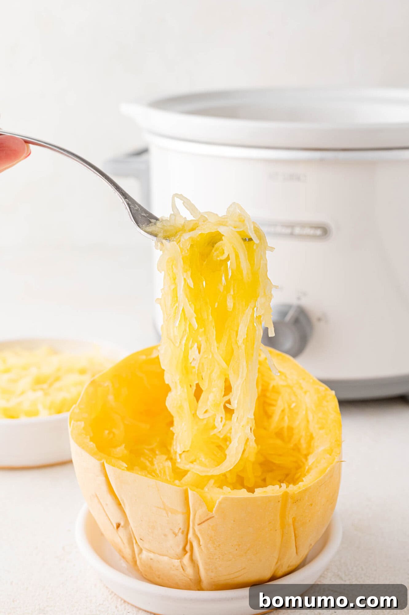 Spaghetti squash strands being lifted out of the squash half with a fork, with a slow cooker in the background.