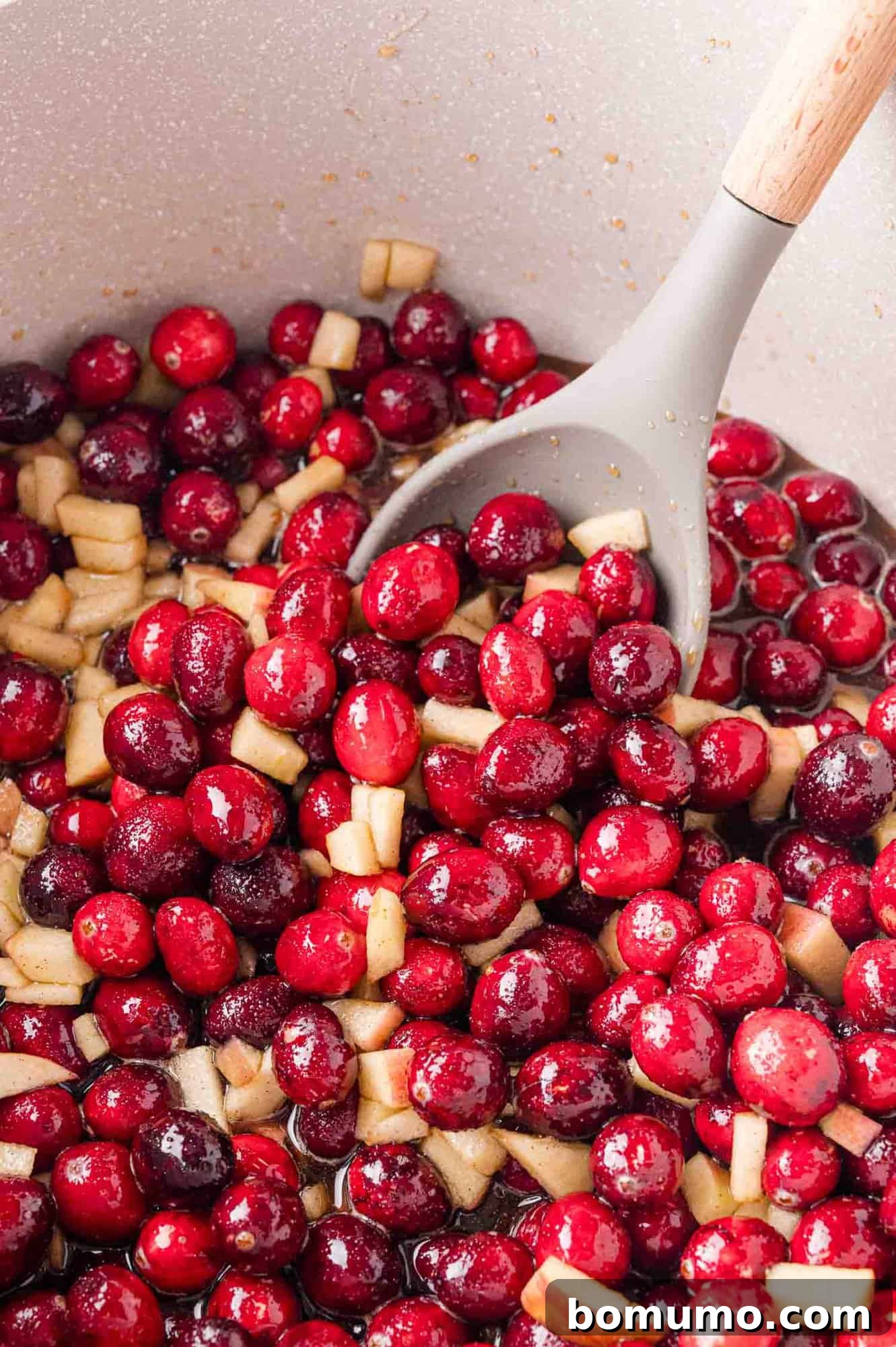 A pot filled with red cranberries and apple pieces, being stirred with a grey spoon. 