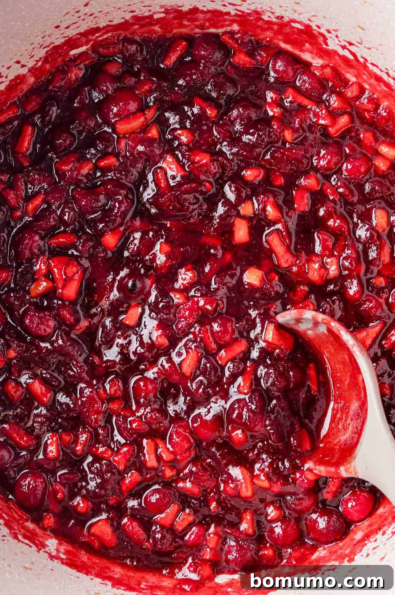 Close-up of a pot filled with chunky cranberry sauce being stirred with a spoon.