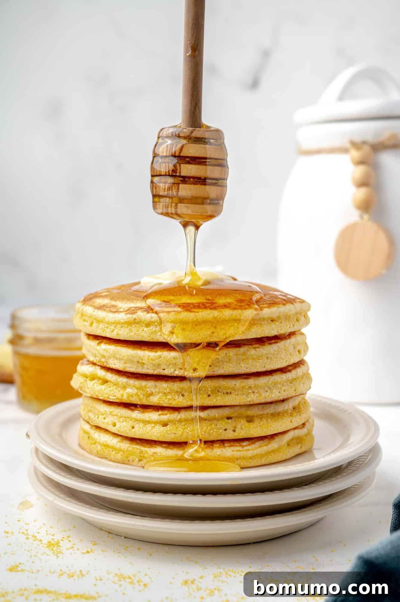 A delectable stack of golden cornbread pancakes, generously drizzled with glistening honey from a wooden dipper. In the soft-focus background, a rustic jar of honey and a pristine white kitchen canister complete the inviting scene.