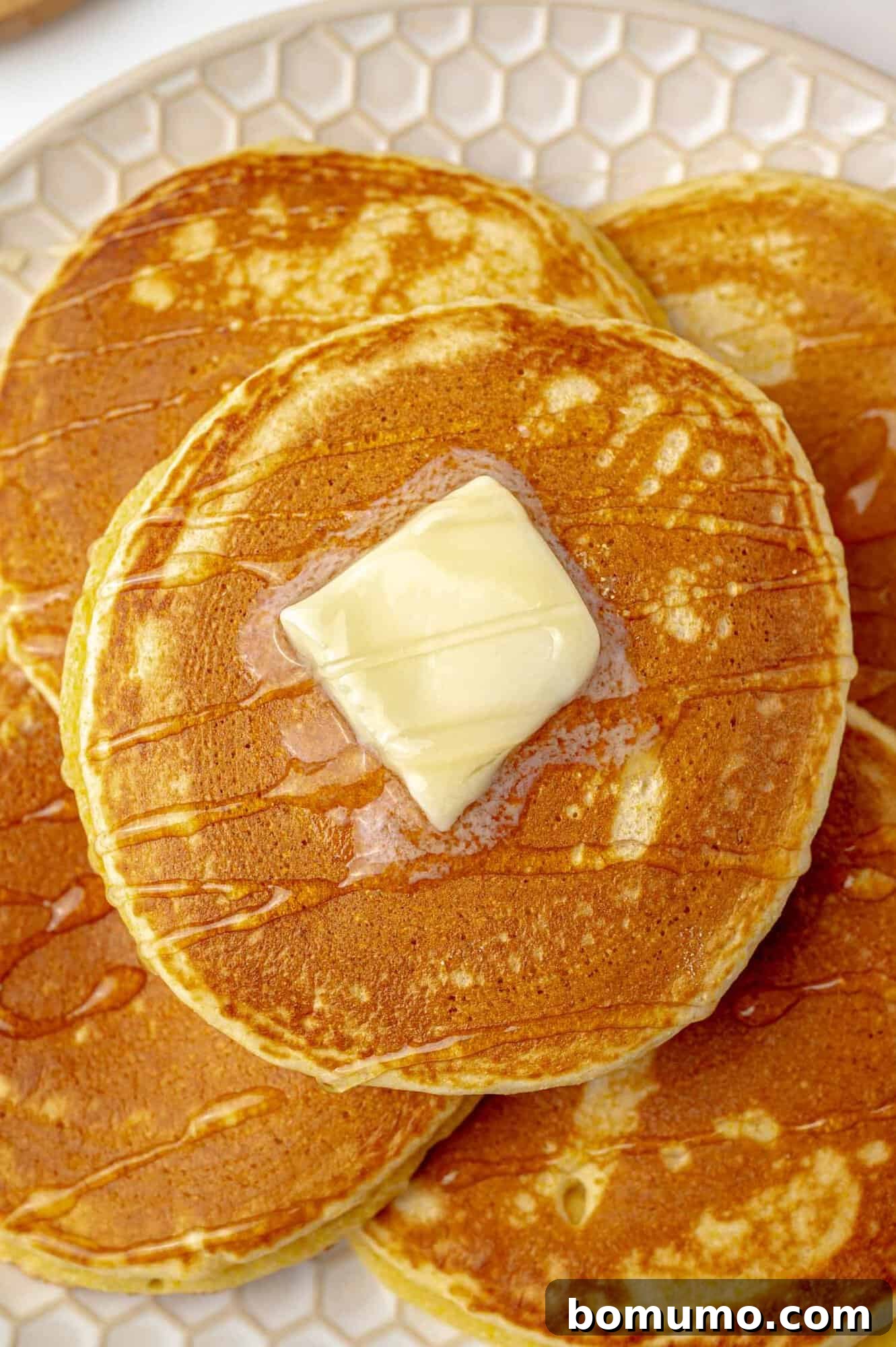 A beautifully stacked tower of golden pancakes, crowned with a melting pat of butter and glistening with sweet syrup, sits invitingly on a charming patterned plate, ready to be enjoyed.