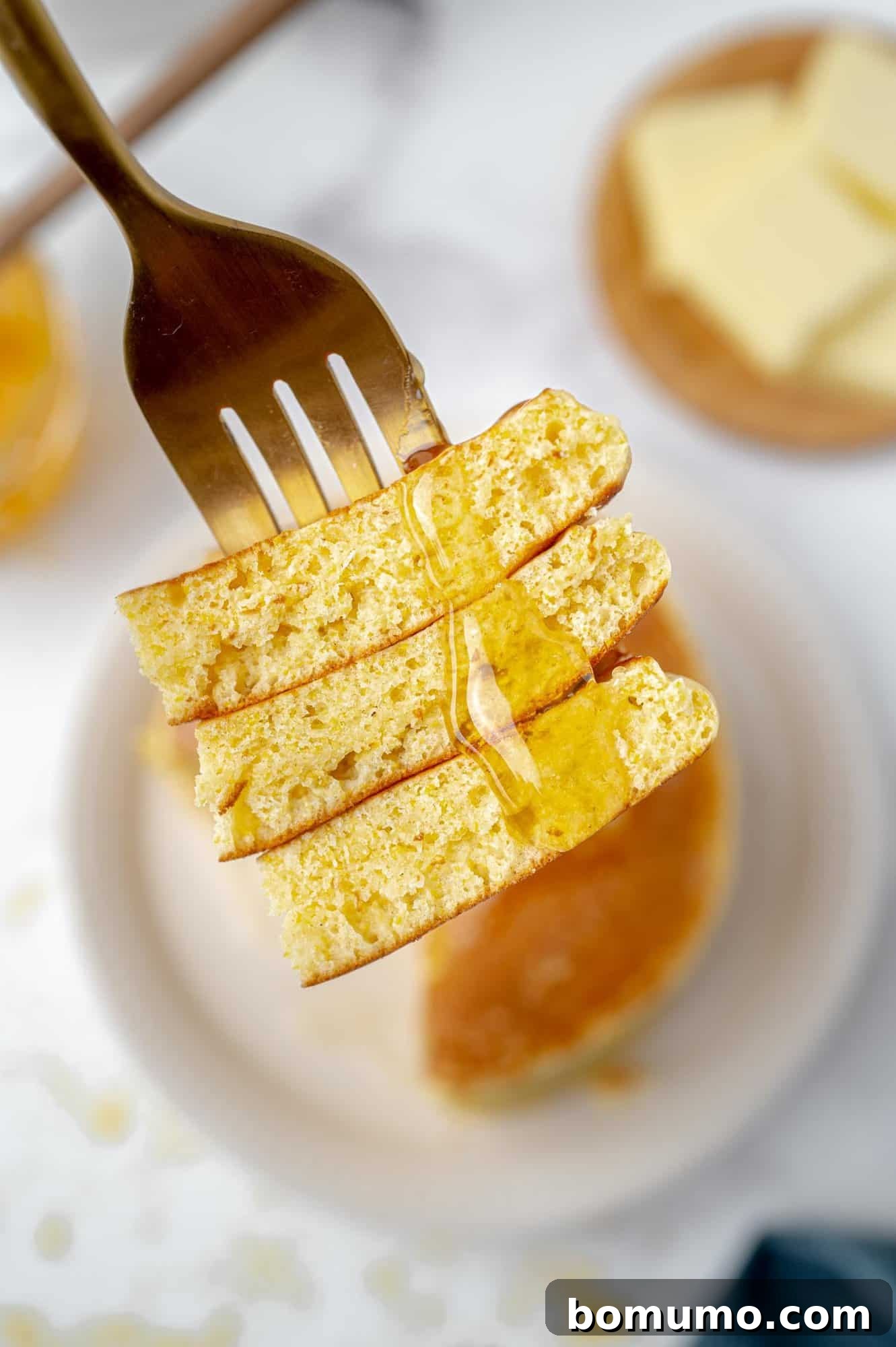 A fork lifts three irresistible, syrup-drizzled pancake pieces, hovering above a plate laden with more golden pancakes. In the inviting background, a charming butter dish awaits, completing this perfect breakfast tableau.