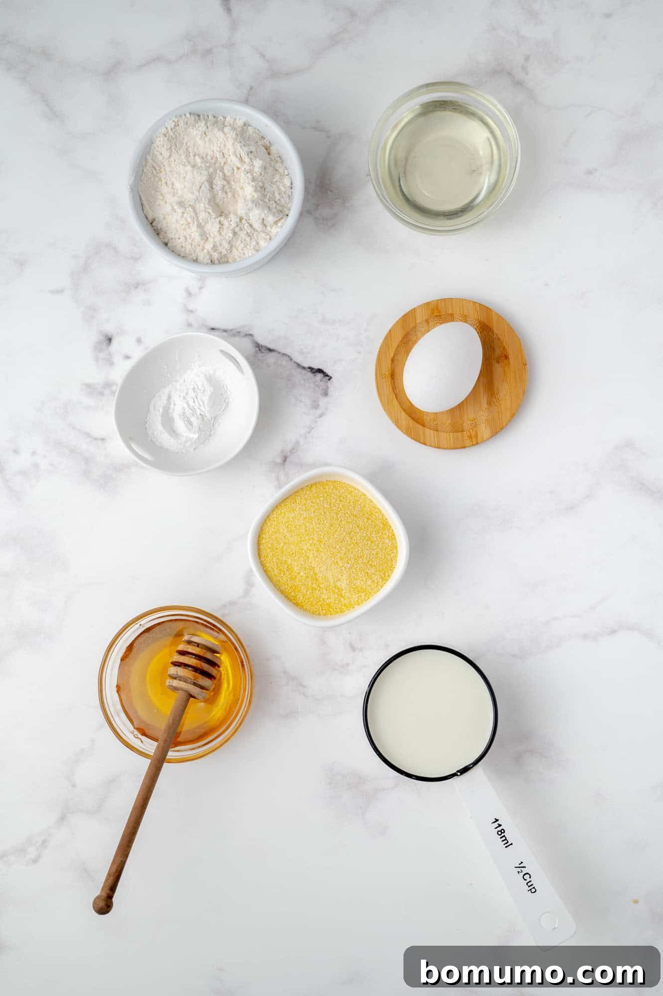 A pristine marble surface showcases the essential ingredients for cornbread pancakes: neatly portioned flour, a measuring cup of oil, baking powder, golden cornmeal, a jar of honey with a dipper, milk in a clear measuring cup, and an empty wooden lid. All arranged for culinary readiness.